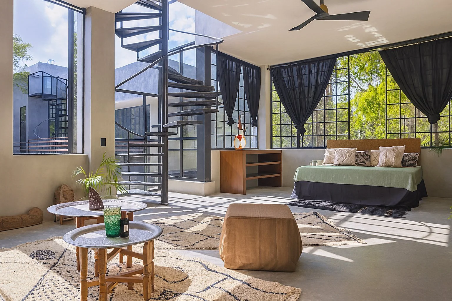 Spacious living room with large windows, spiral staircase, black curtains, bed with multiple pillows, and modern decor.