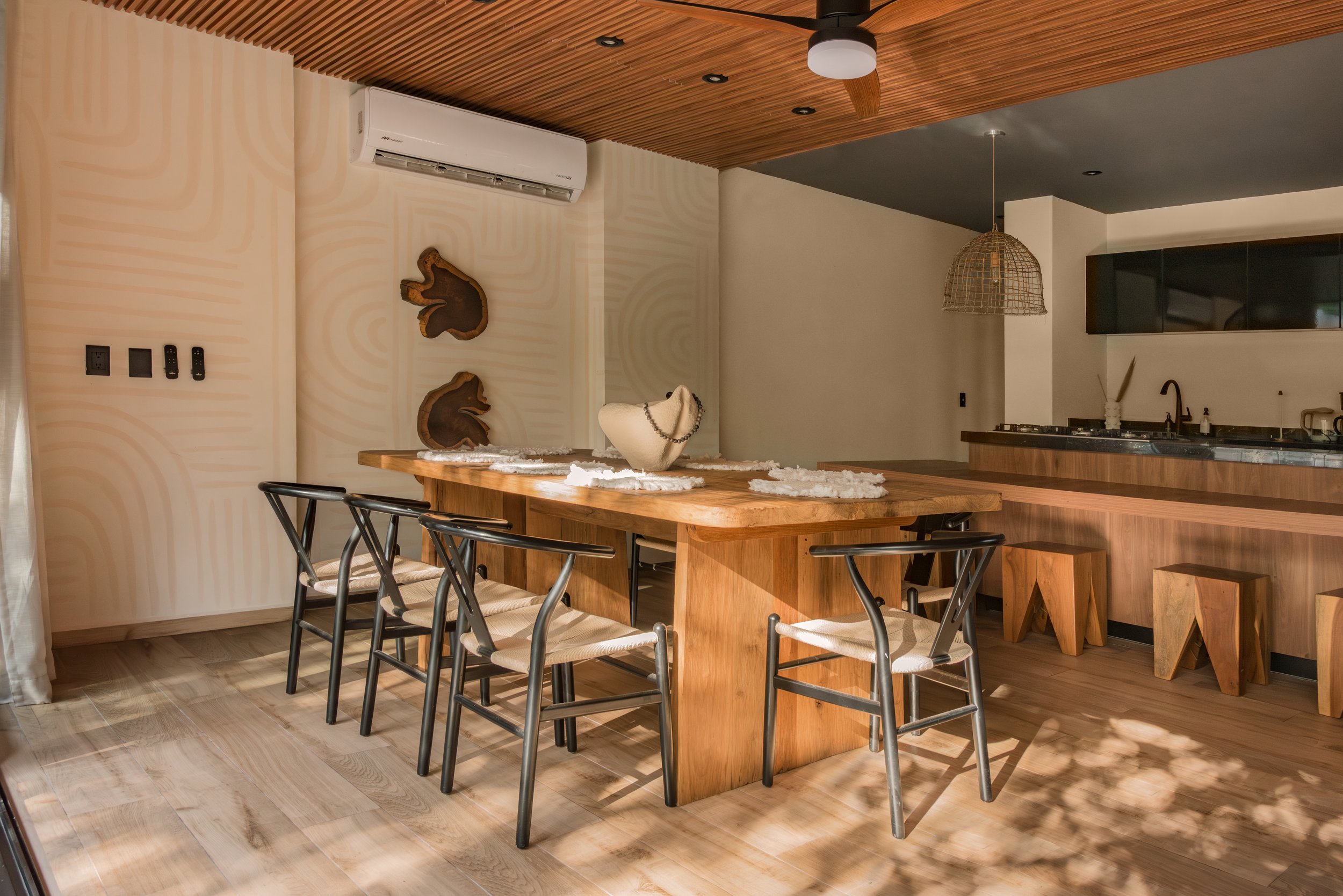 A wooden dining table surrounded by black chairs in a modern kitchen and dining area with beige walls, wooden ceiling, and decorative wood art on the wall.
