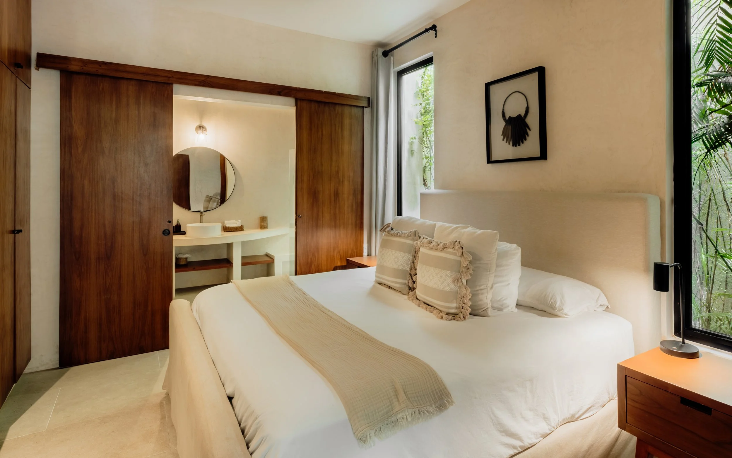 A bedroom featuring a large bed with white linens, decorative pillows, and a throw blanket. There are wooden sliding doors, a window with curtains, and a framed black artwork on the wall. A small wooden nightstand with a modern black lamp is next to the bed. The room has a natural and minimalist aesthetic.