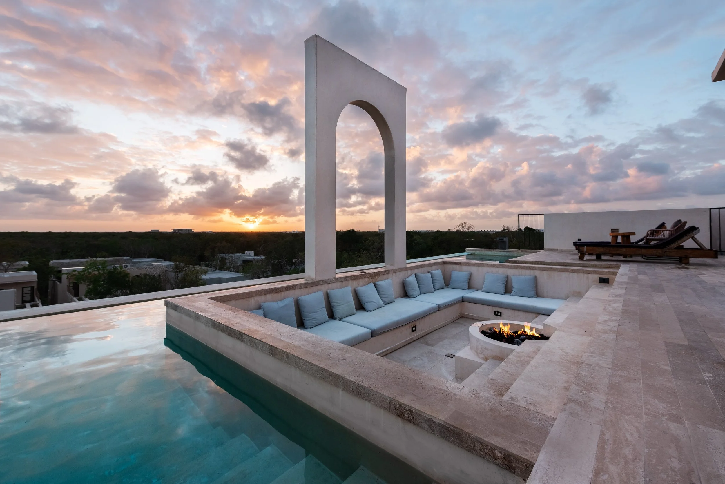 A luxurious rooftop pool area at sunset with comfortable white cushioned seating, a fire pit, a fire feature, and a scenic view of the sunset and cloudy sky.