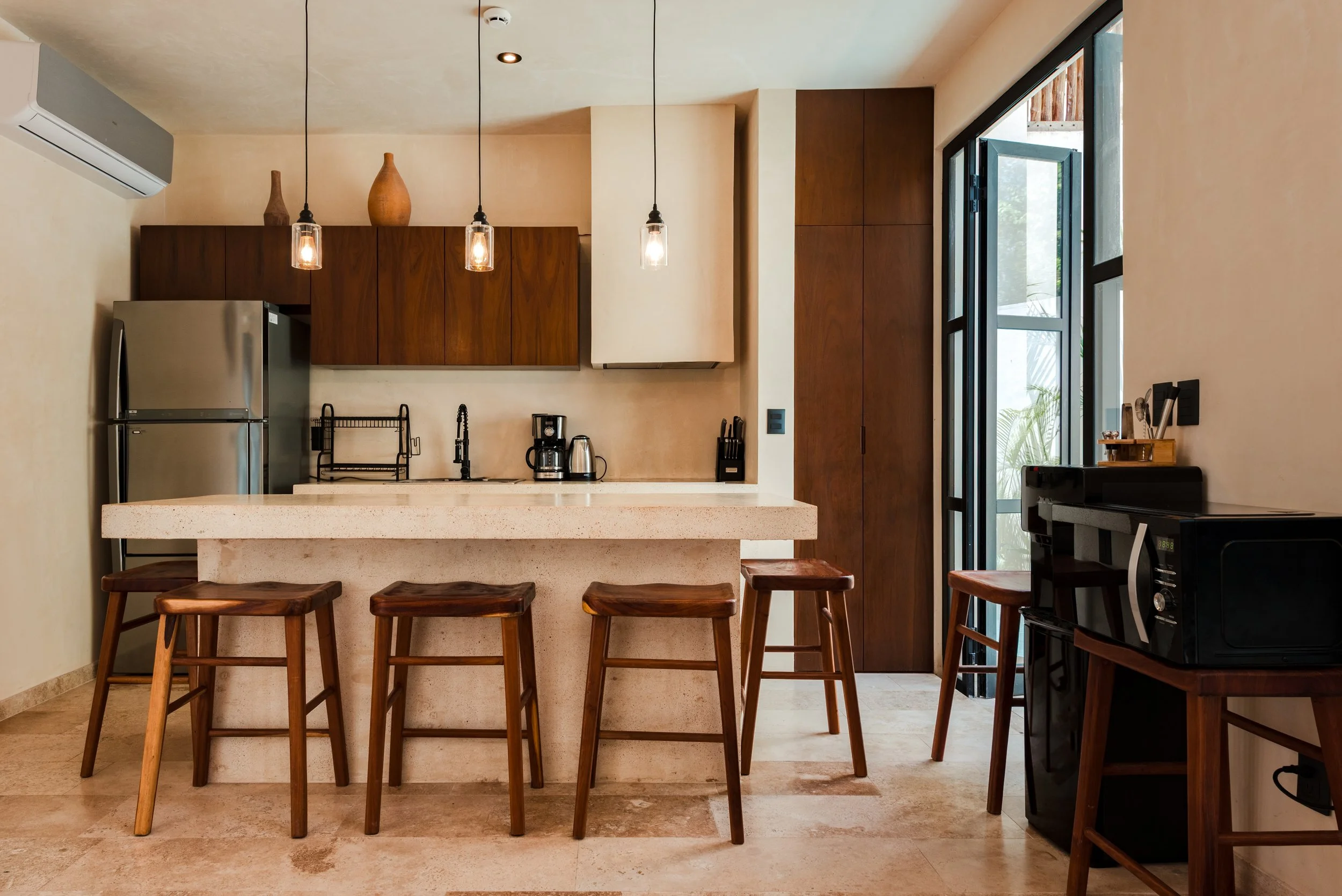 Modern kitchen with brown wooden cabinets, stainless steel refrigerator, black sink, coffee maker, toaster, pendant lights, and a large window with black frames.