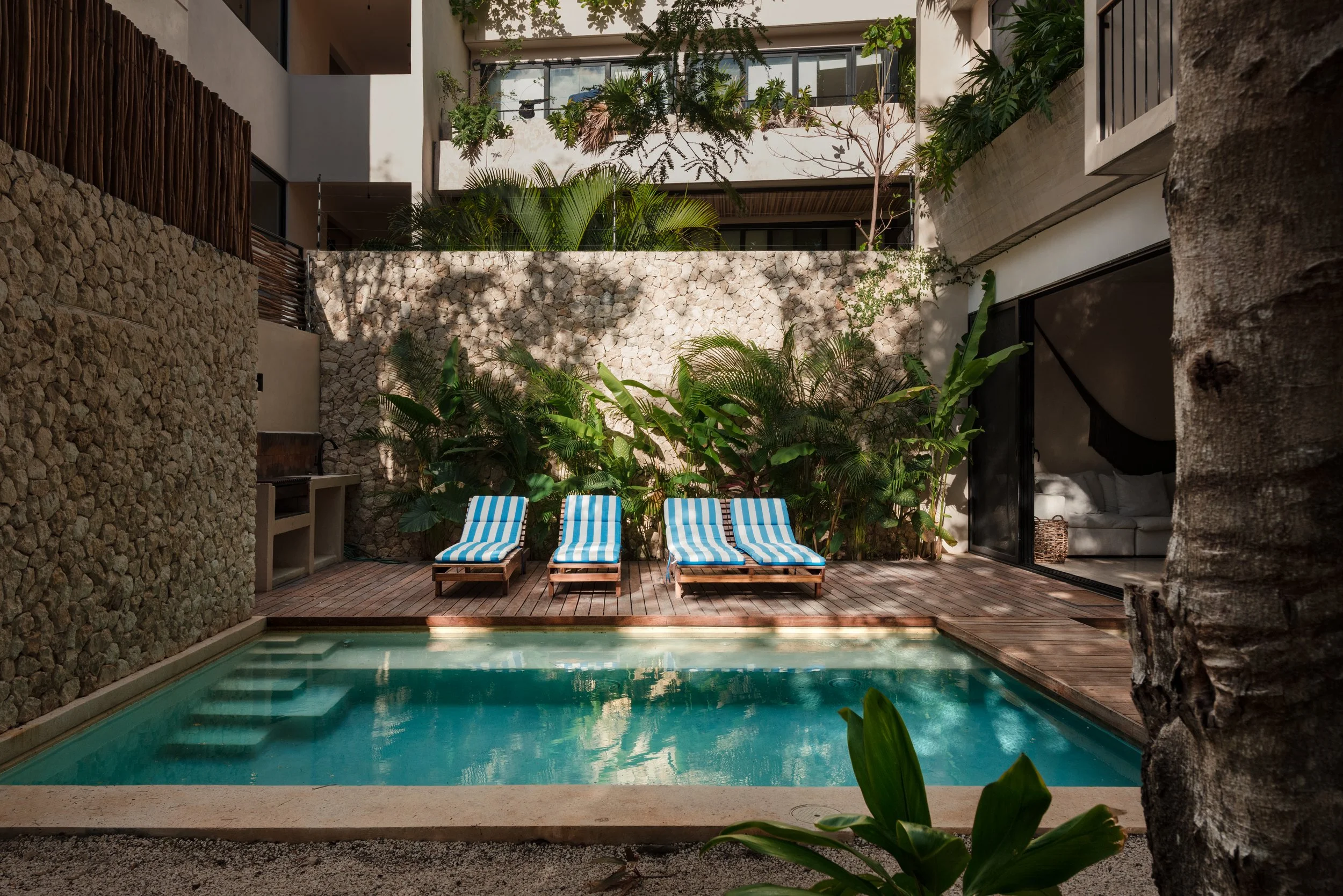 A backyard pool with three blue and white striped lounge chairs on a wooden deck, surrounded by lush greenery and a stone wall, with modern apartment buildings in the background.