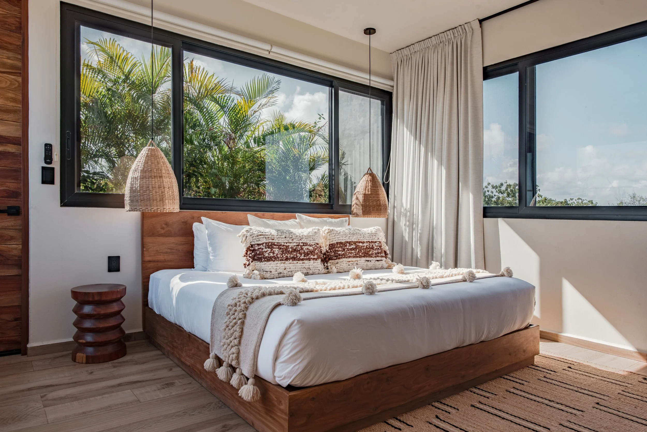 A bedroom with a wooden bed frame, white bedding, decorative pillows, and a throw blanket with tassels. There are large windows with black frames and white curtains, revealing green tropical foliage outside. Wicker pendant lights hang on either side of the bed, and a wooden side table is on the left. The room has wood flooring and a patterned rug.