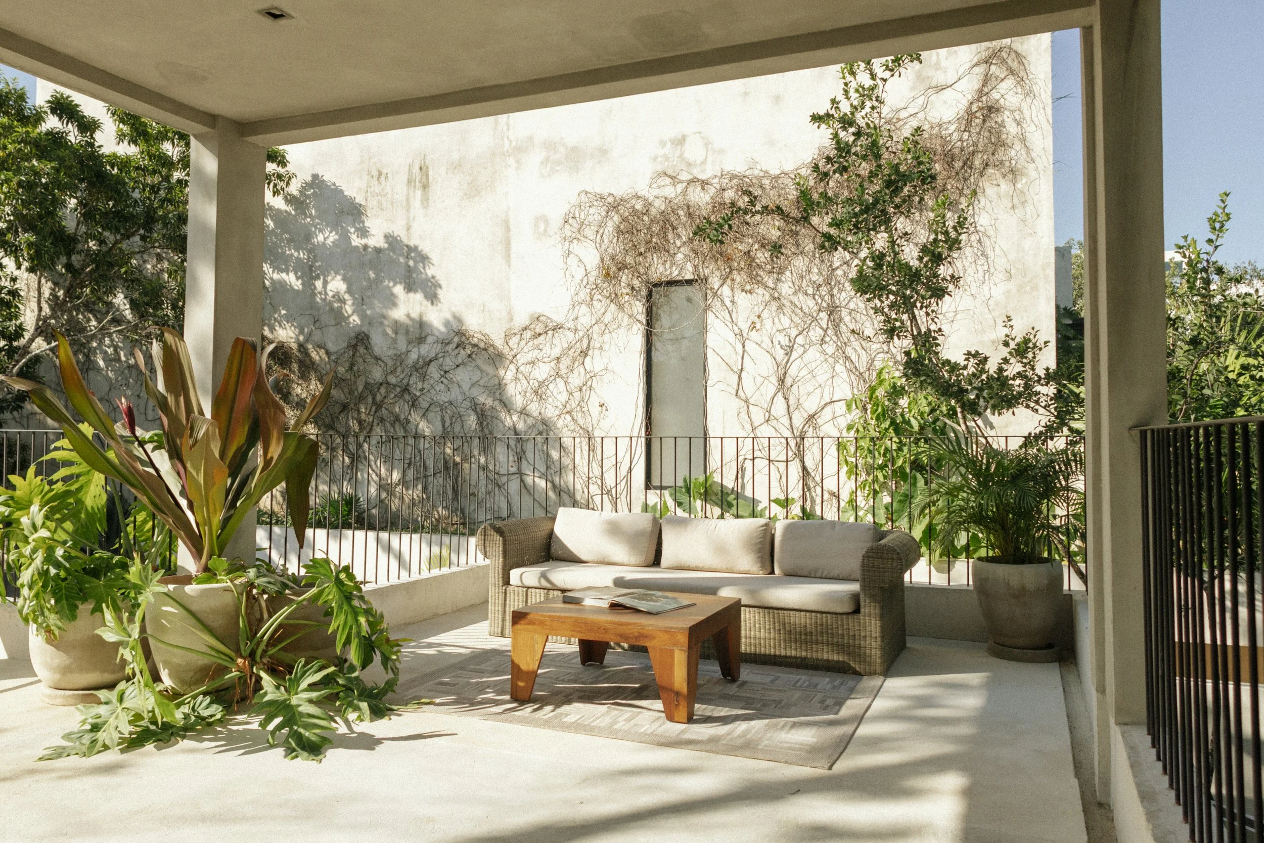 Outdoor patio with a wicker sofa and a wooden coffee table, surrounded by potted plants and greenery, with a white wall and trees in the background.