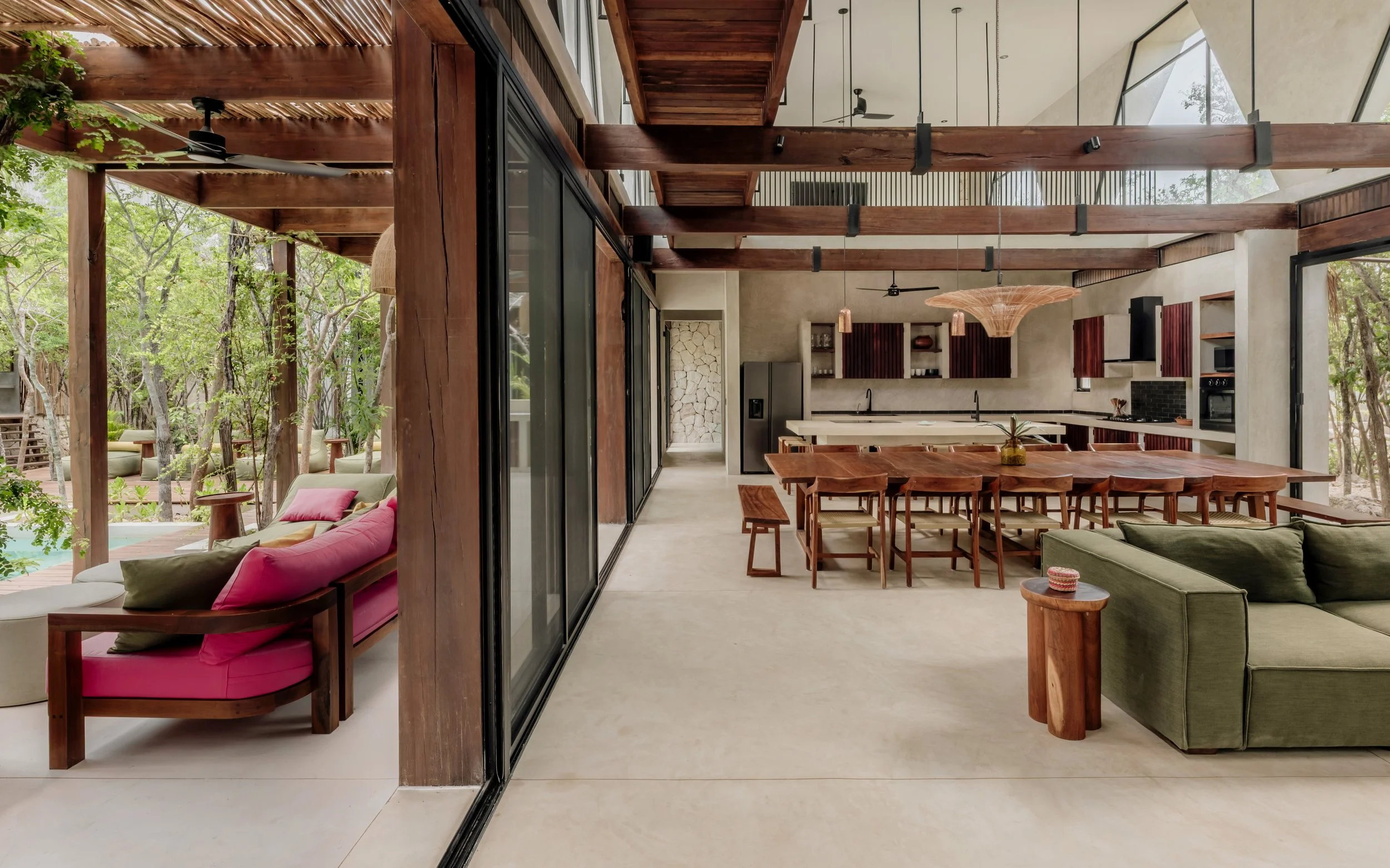 Open-concept living and dining area with wood beams, modern kitchen, and large glass sliding doors leading to outdoor patio with seating and lush trees.