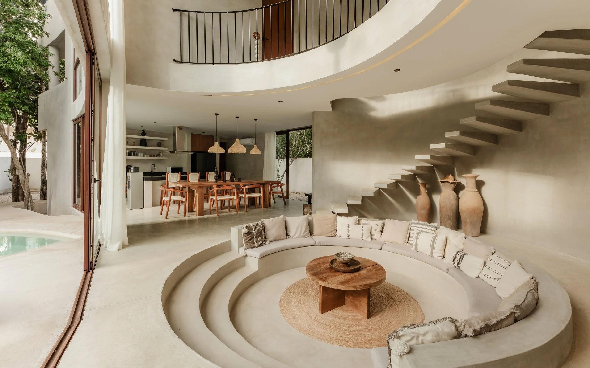 Open-plan living and dining area with curved beige sofa, round wooden coffee table, staircase with concrete steps, dining table with wooden chairs, industrial-style pendant lights, and large glass doors leading outside.