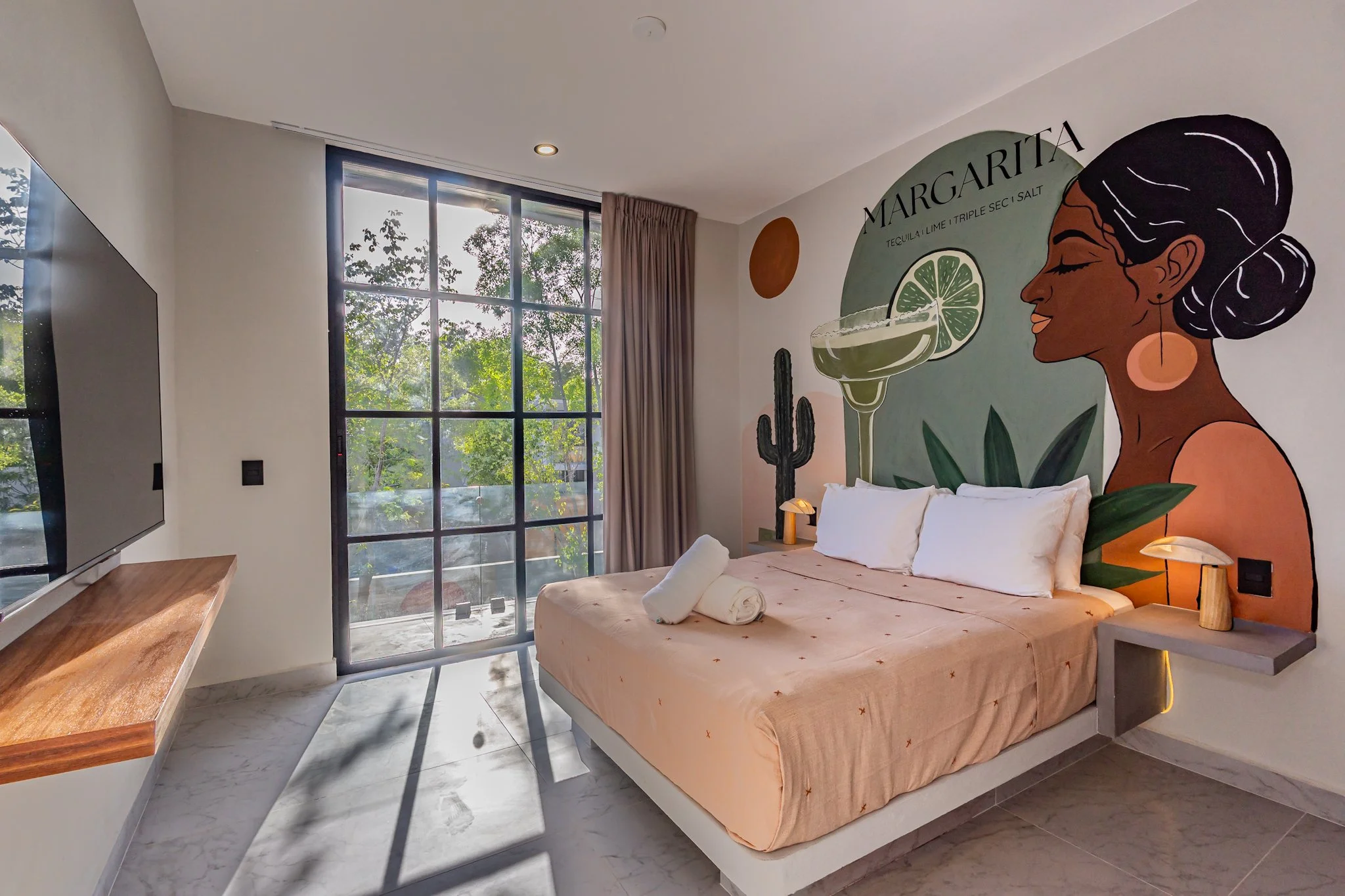 A hotel room featuring a large window with trees outside, a mural of a woman with dark hair and a large earring, and a bed with folded towels. The mural advertises a margarita drink.