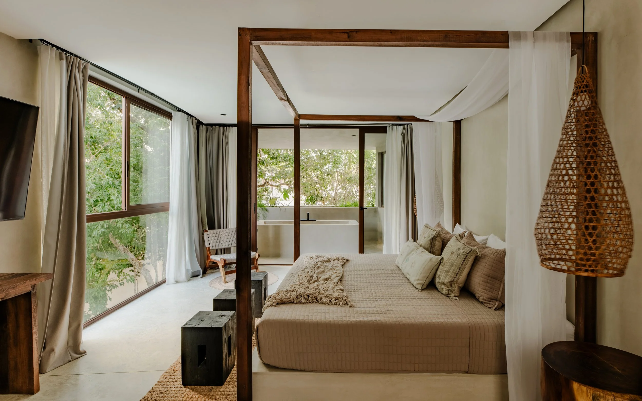 A cozy modern bedroom with large windows overlooking green trees, a beige bed with multiple pillows, a woven hanging lamp, and a wooden frame separating the sleeping area from the balcony.