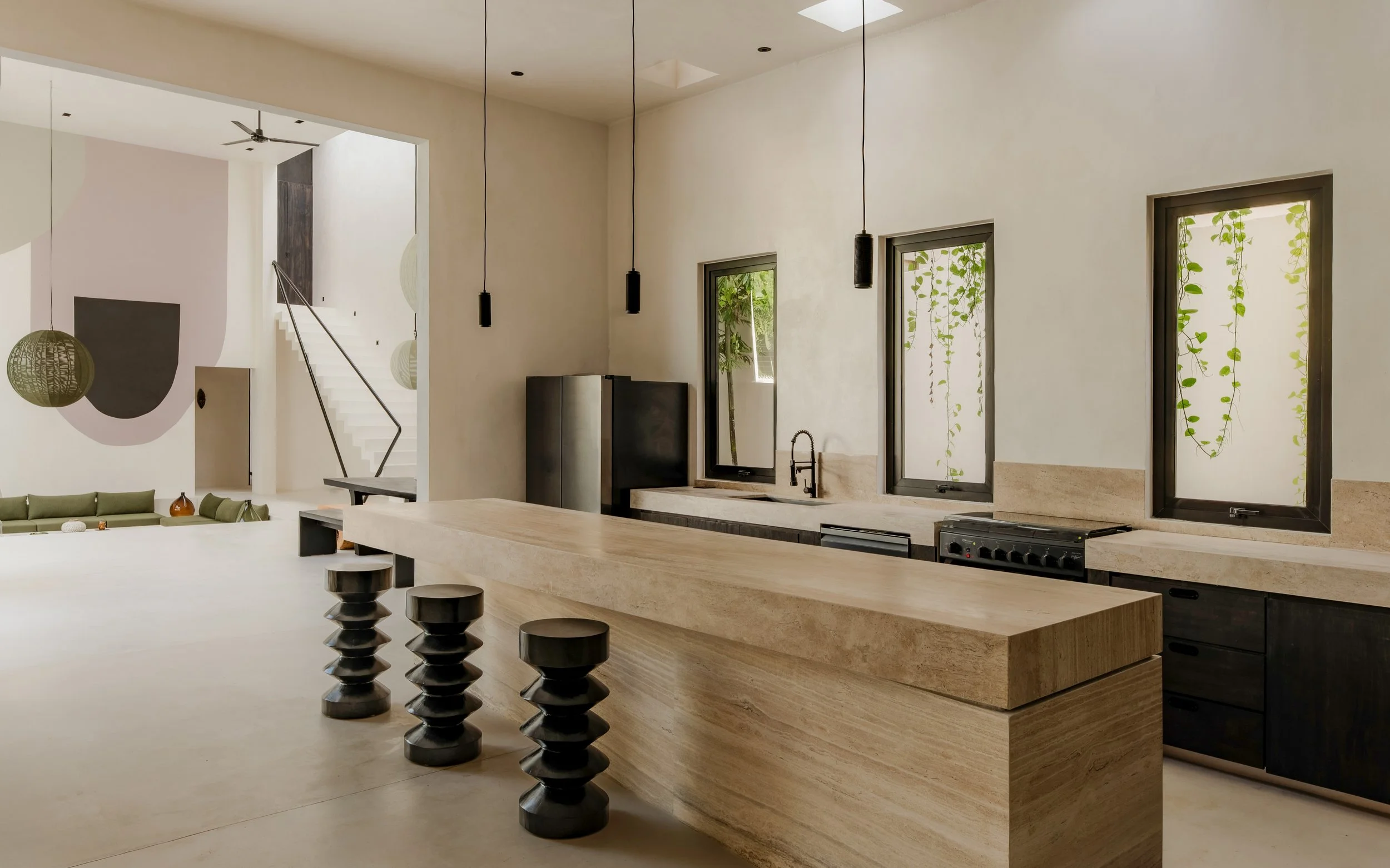Modern minimalist kitchen with beige stone countertop, black pendant lights, large windows with greenery outside, and black stools at the island.