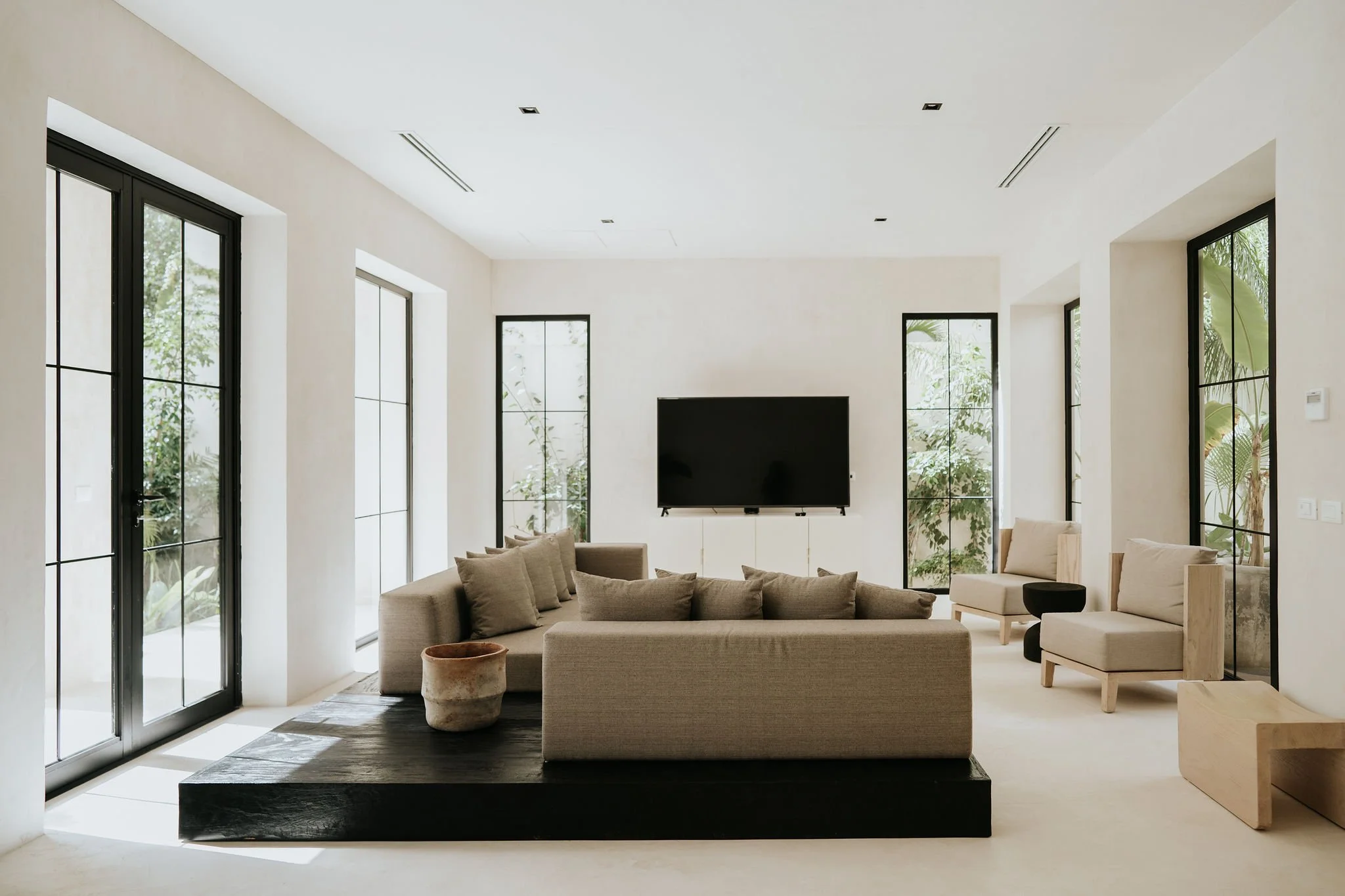 Bright living room with large windows, beige sofas, a black TV on a white cabinet, and minimal decor.