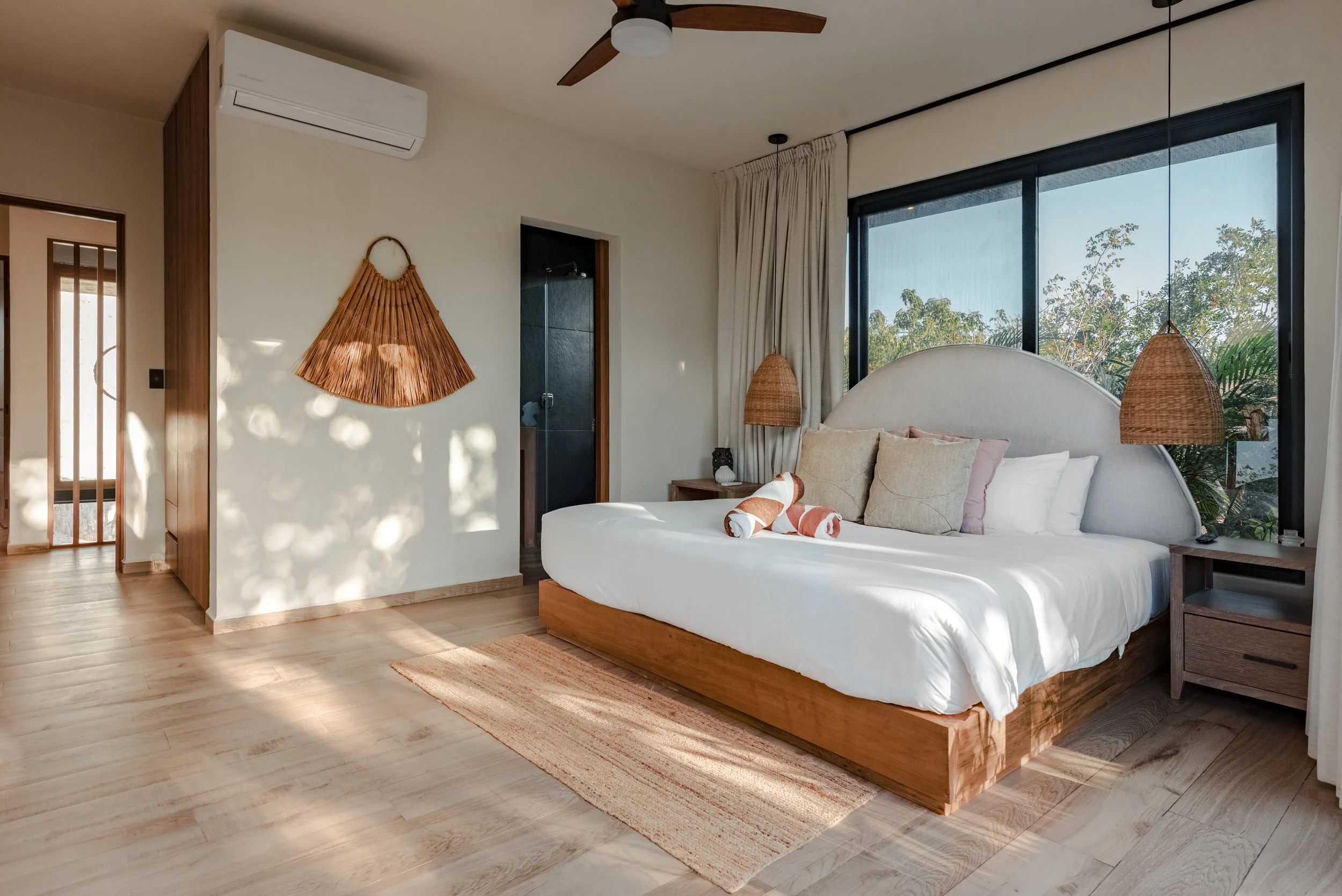 A bedroom with a large bed, beige headboard, and rolled towels on the white bedding. There are two rattan pendant lights hanging on either side of the bed, beige curtains, and a large window showing trees outside. Light wood flooring and a ceiling fan are also visible.