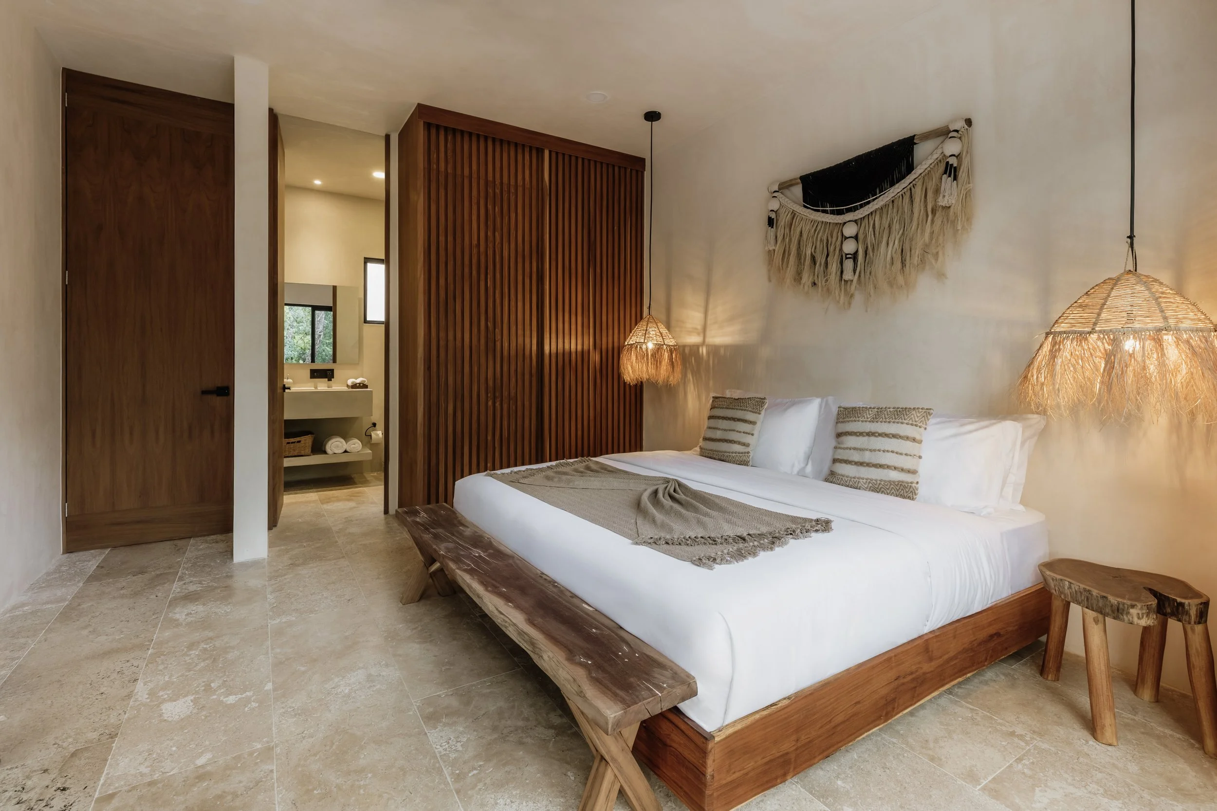 A cozy bedroom with a white bed and beige pillows, decorated with a bohemian wall hanging and pendant lights, with an attached bathroom visible through an open doorway.