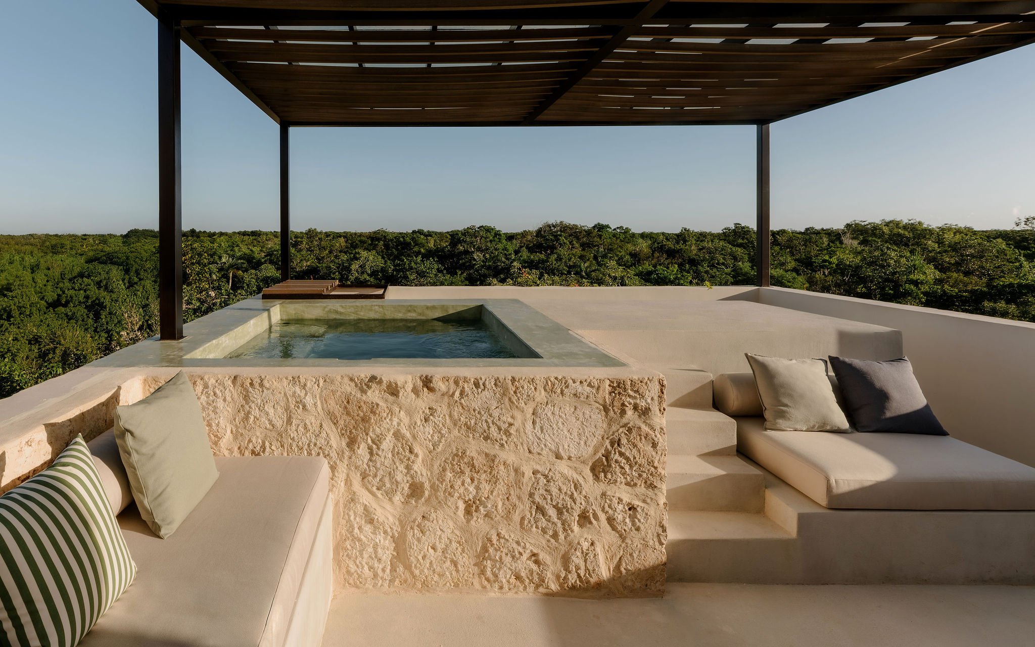 A modern outdoor patio with a built-in hot tub, cushioned lounge seating, and a view of lush green treetops under a wooden pergola.
