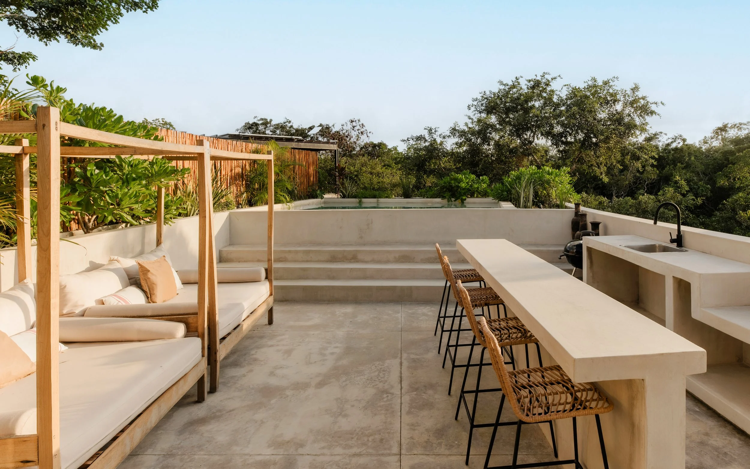 Outdoor rooftop patio with white seating area, bar counter with barstools, lush green trees and blue sky.