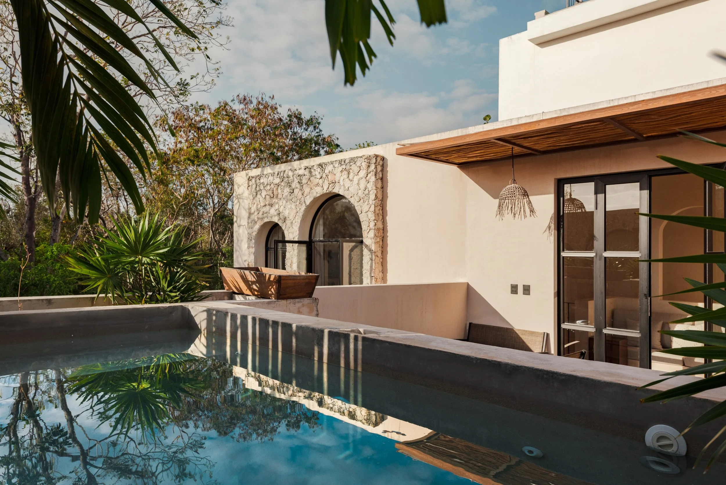 Modern house with stone and white walls, large sliding glass doors, wooden pergola, lush green plants, and reflecting pool under a partly cloudy sky.