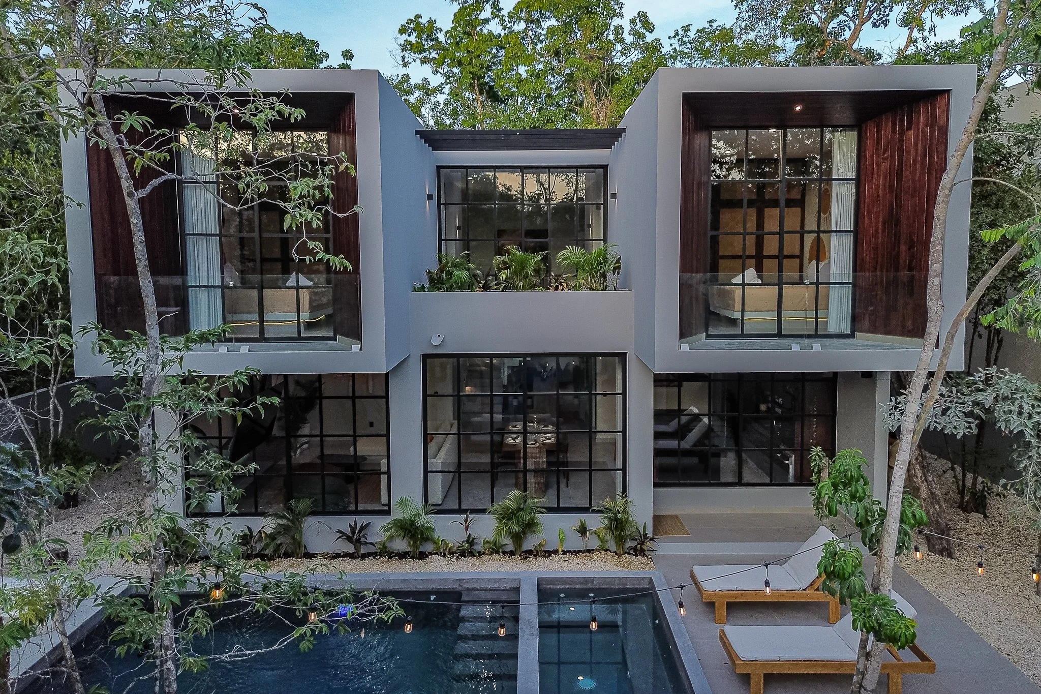 Modern multi-story house with large glass windows, surrounded by trees and plants, with a swimming pool and outdoor seating area.