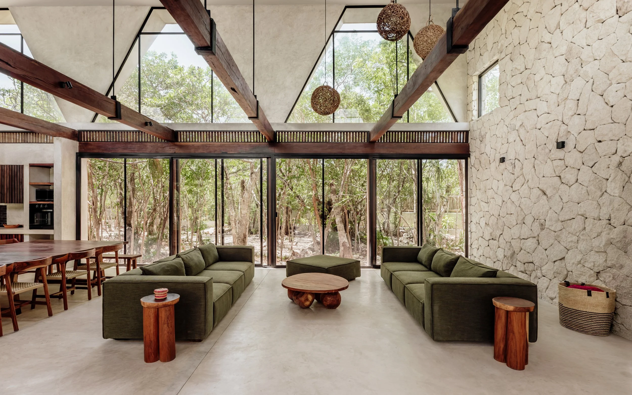 Modern living room with large windows, stone wall, wooden beams, and green sectional sofa, overlooking a wooded outdoor area.