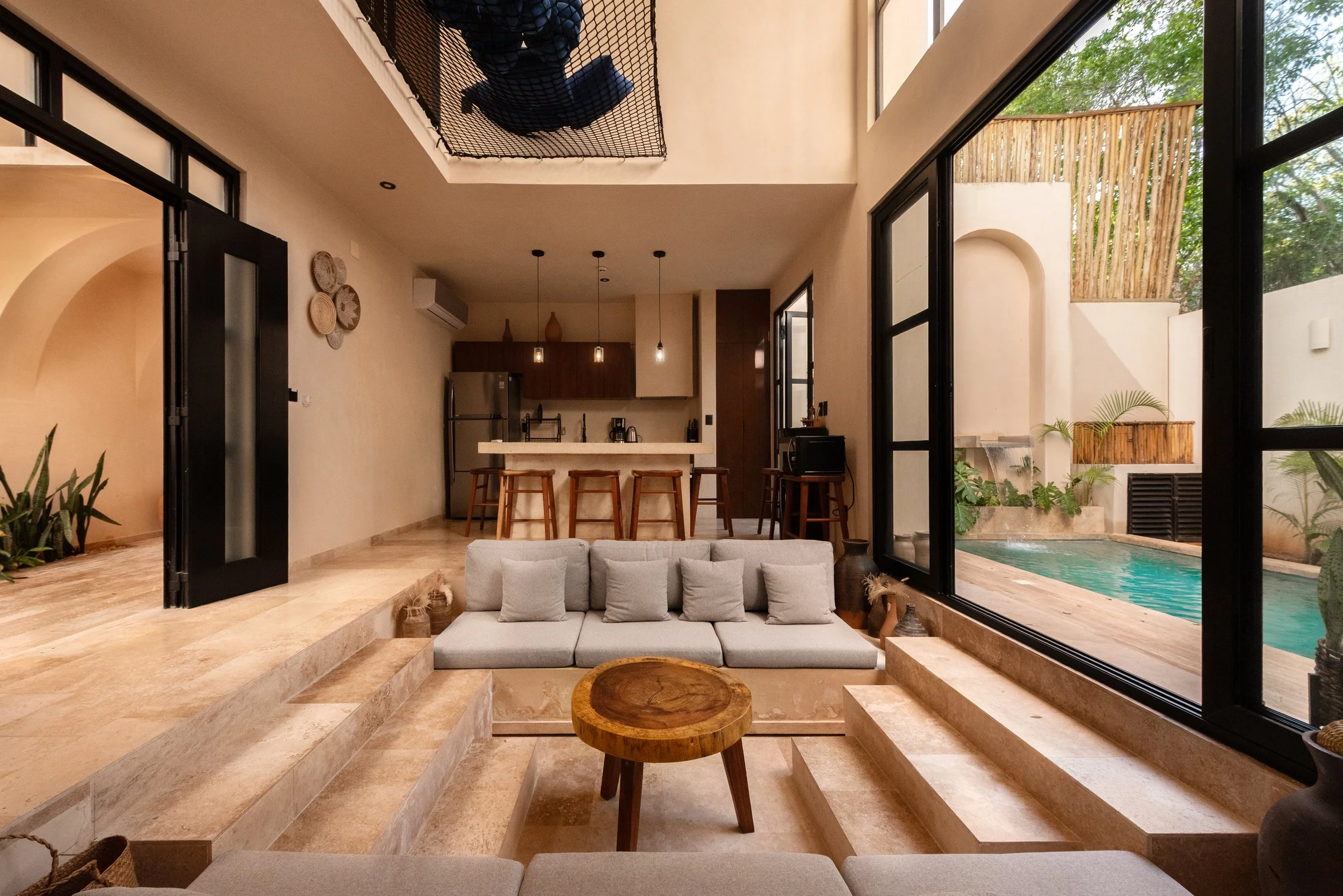 Living room with large open windows overlooking a pool and outdoor space, beige tiled flooring, a white couch with pillows, and a kitchen in the background.
