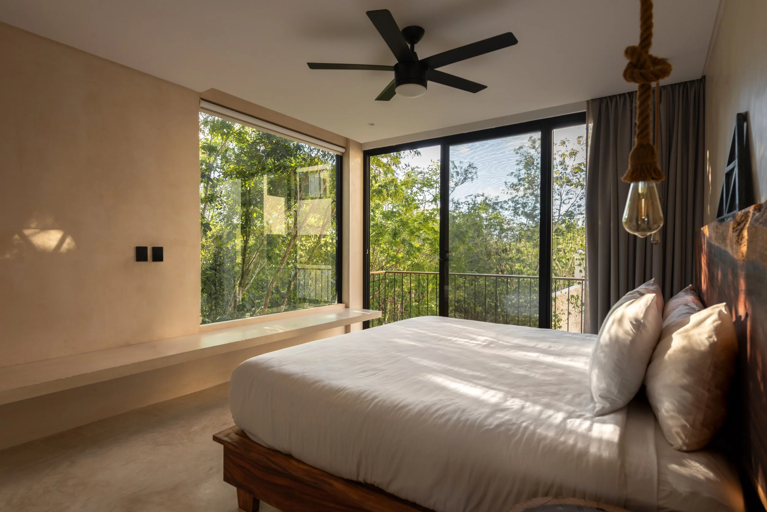 A modern bedroom with a bed, large glass windows showing trees outside, ceiling fan, and a hanging light bulb.