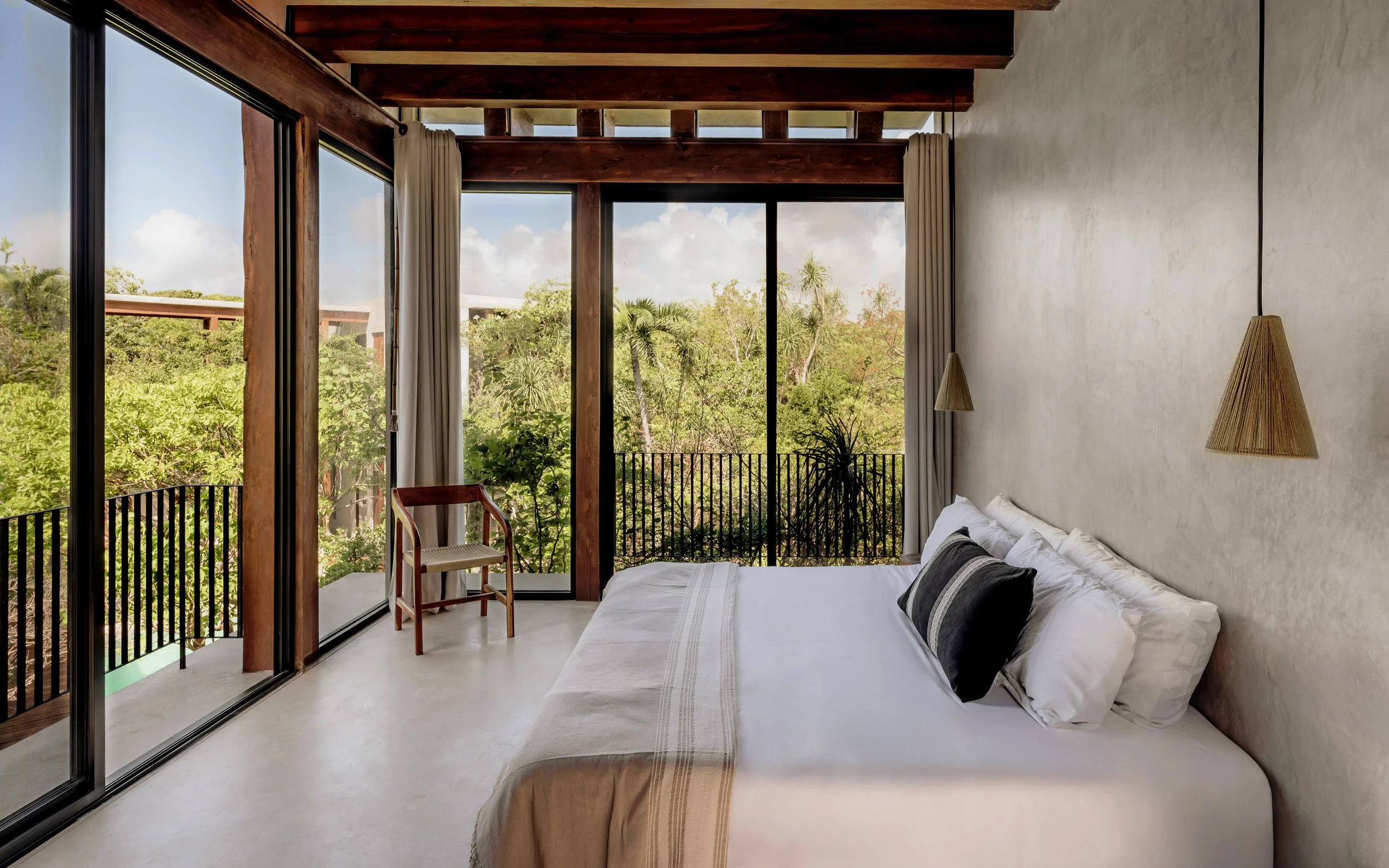 Bedroom with a large window showing greenery outside, a bed with white and black pillows, pendant lights, and a wooden chair on the balcony.