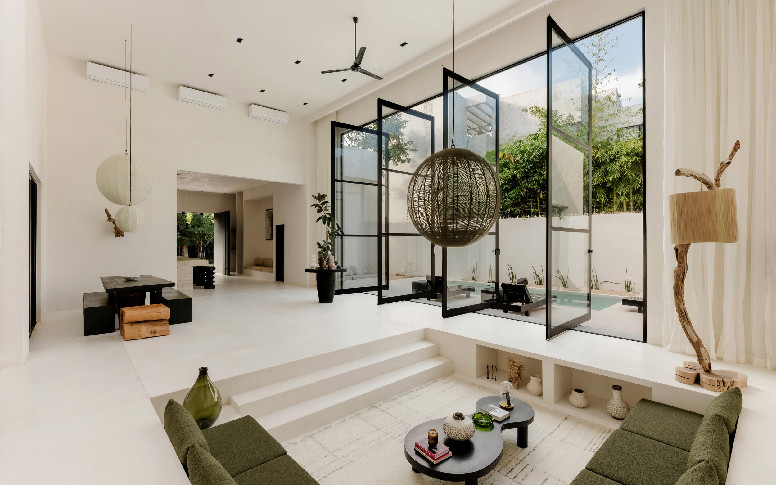 Modern living room with white walls, large glass windows, black framework, green sofas, round black coffee table with decorative items, hanging paper lanterns, and a view of outdoor greenery.