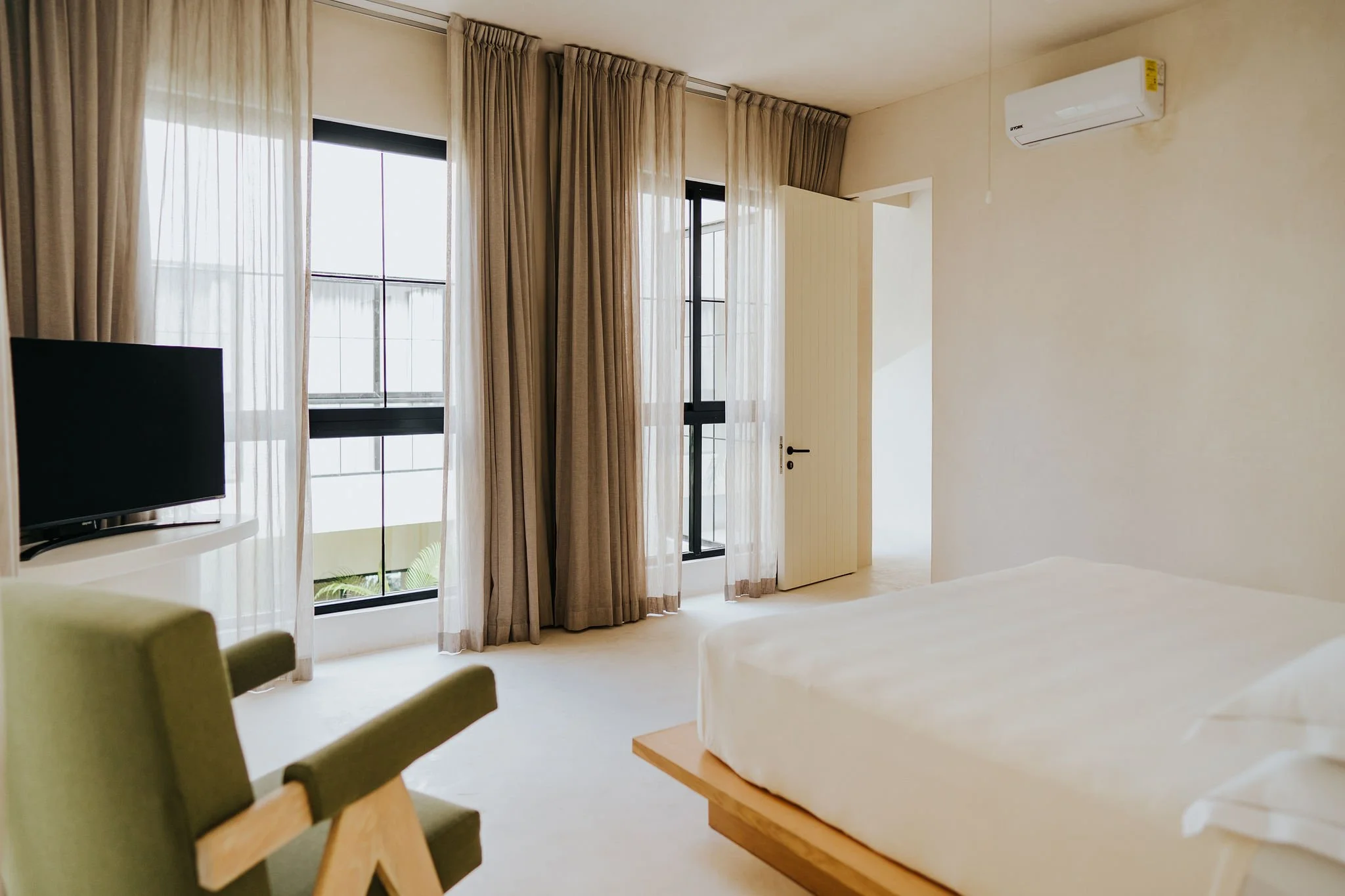 An interior view of a bright hotel room with large windows covered by sheer and beige curtains, a flat-screen TV, a green armchair, a bed with white linens, and an air conditioning unit on the wall.