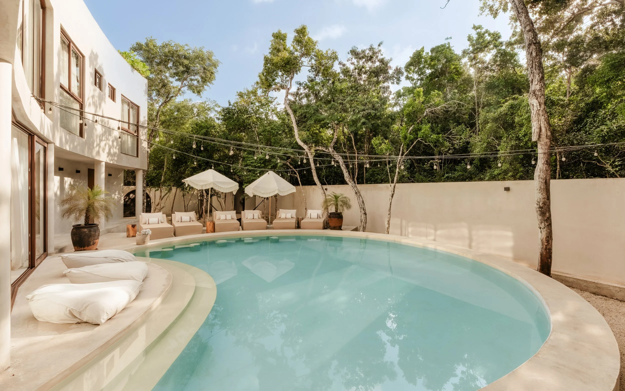 A modern backyard pool area with lounge chairs and umbrellas surrounded by trees and string lights.