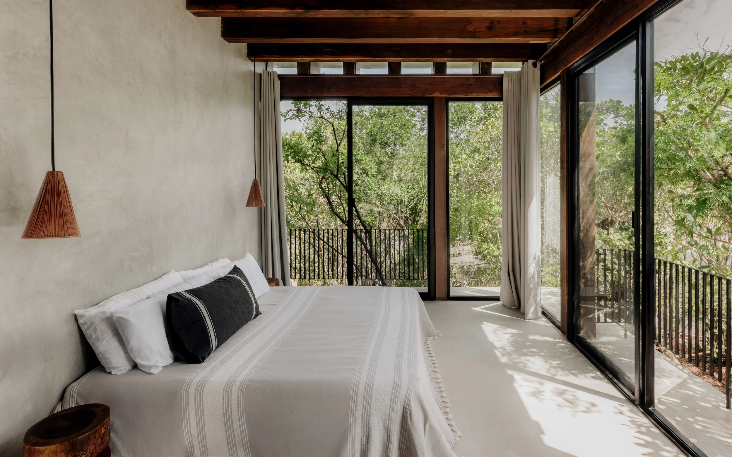 A bedroom with a large bed dressed in white linens, featuring black and white pillows. The room has a modern, minimalist style with a textured white wall and hanging pendant lights with wooden shades. Floor-to-ceiling glass sliding doors open to a balcony with a black railing, and offer a view of green trees outside, allowing natural light to fill the space.