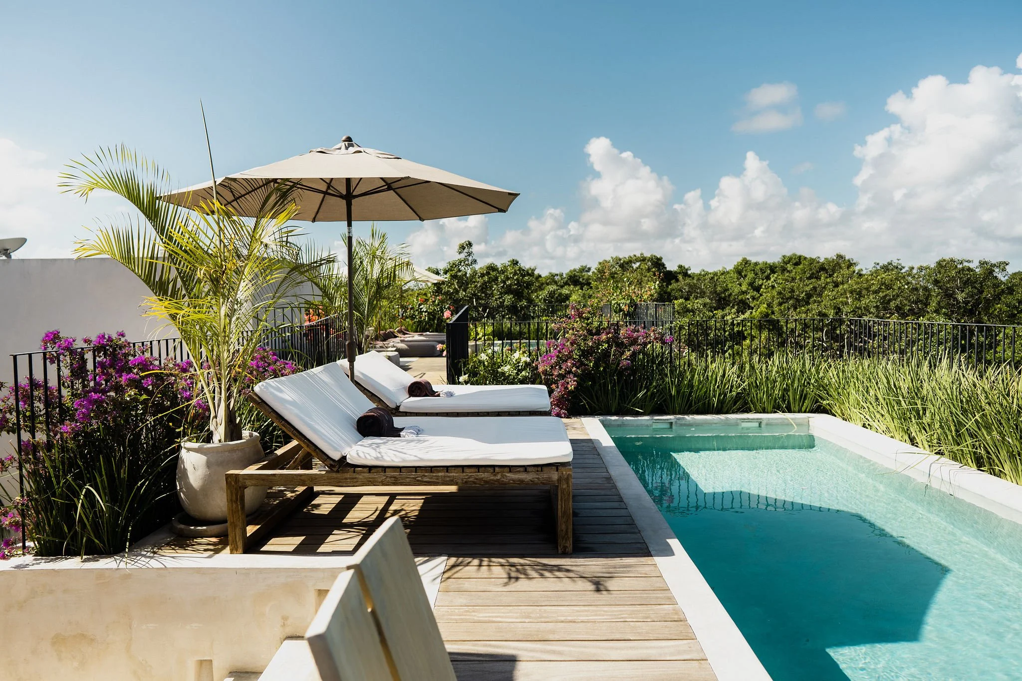 A sunny outdoor poolside patio with white lounge chairs, large potted plants, colorful flowers, a beige umbrella, and a view of trees and a partly cloudy sky.