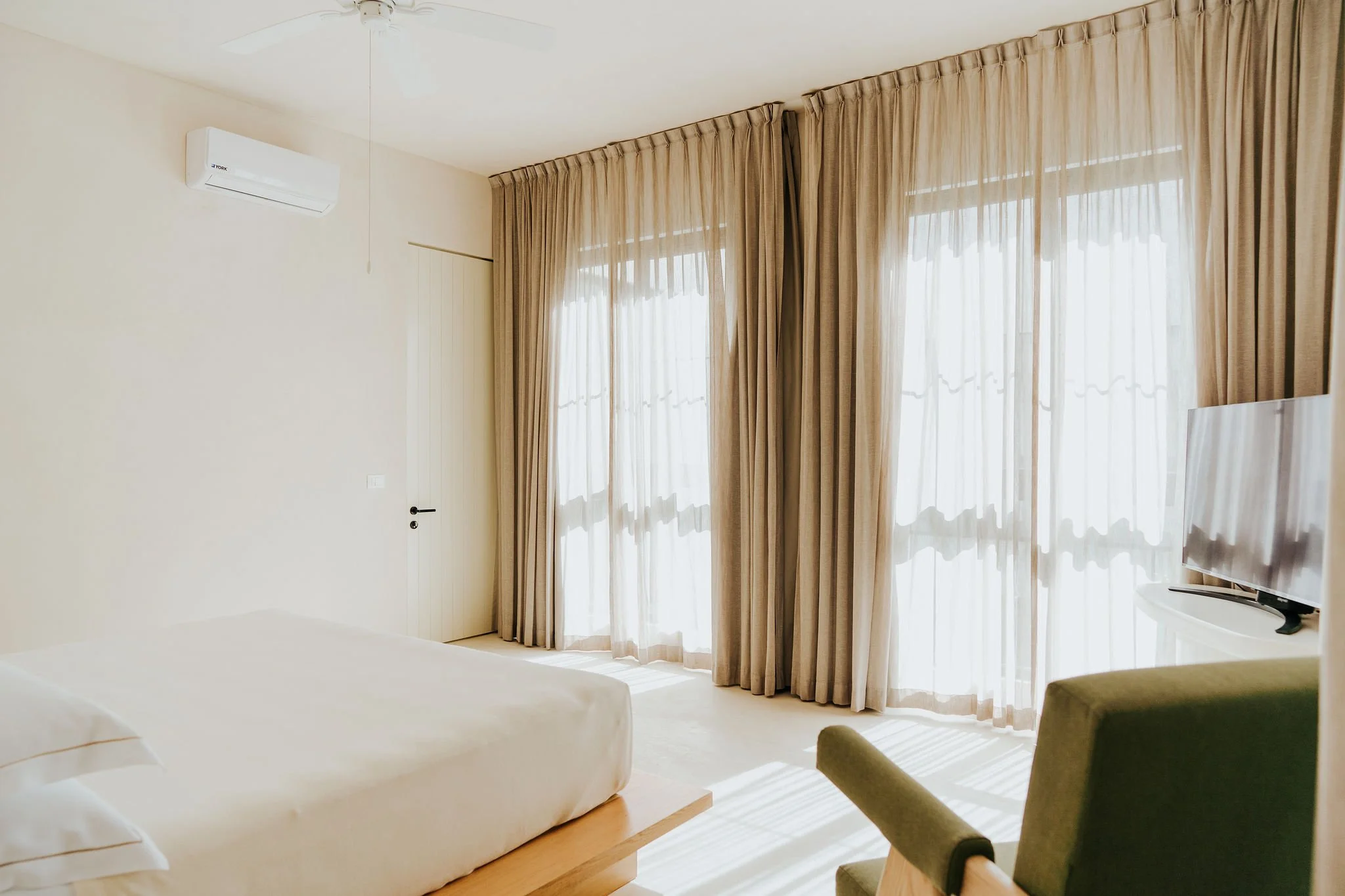 A bright hotel room with large sheer curtains covering floor-to-ceiling windows, a mounted air conditioner on the wall, a bed with white bedding, a green armchair, and a flat-screen TV.