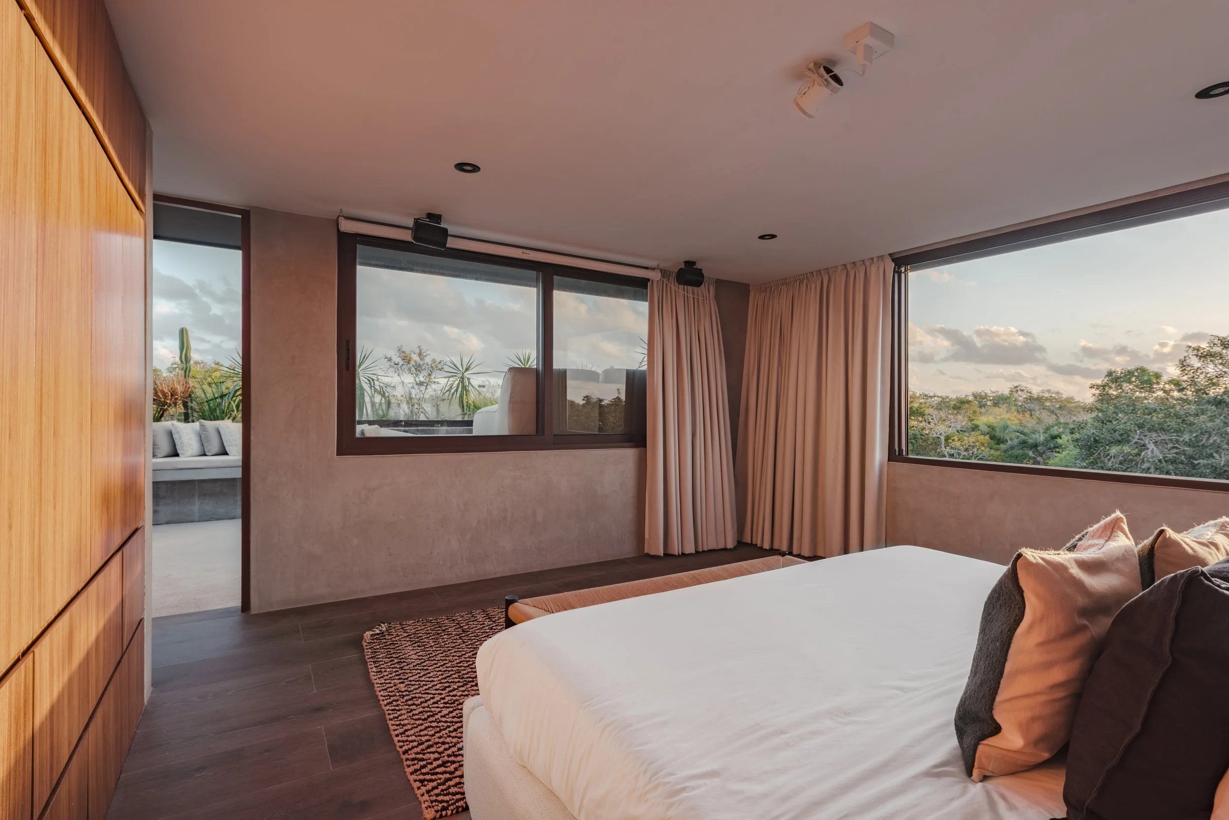 A bedroom with large windows showing outdoor trees and sky, cream-colored curtains, a bed with brown pillows, a zebra-print rug, and wood-paneled wall.