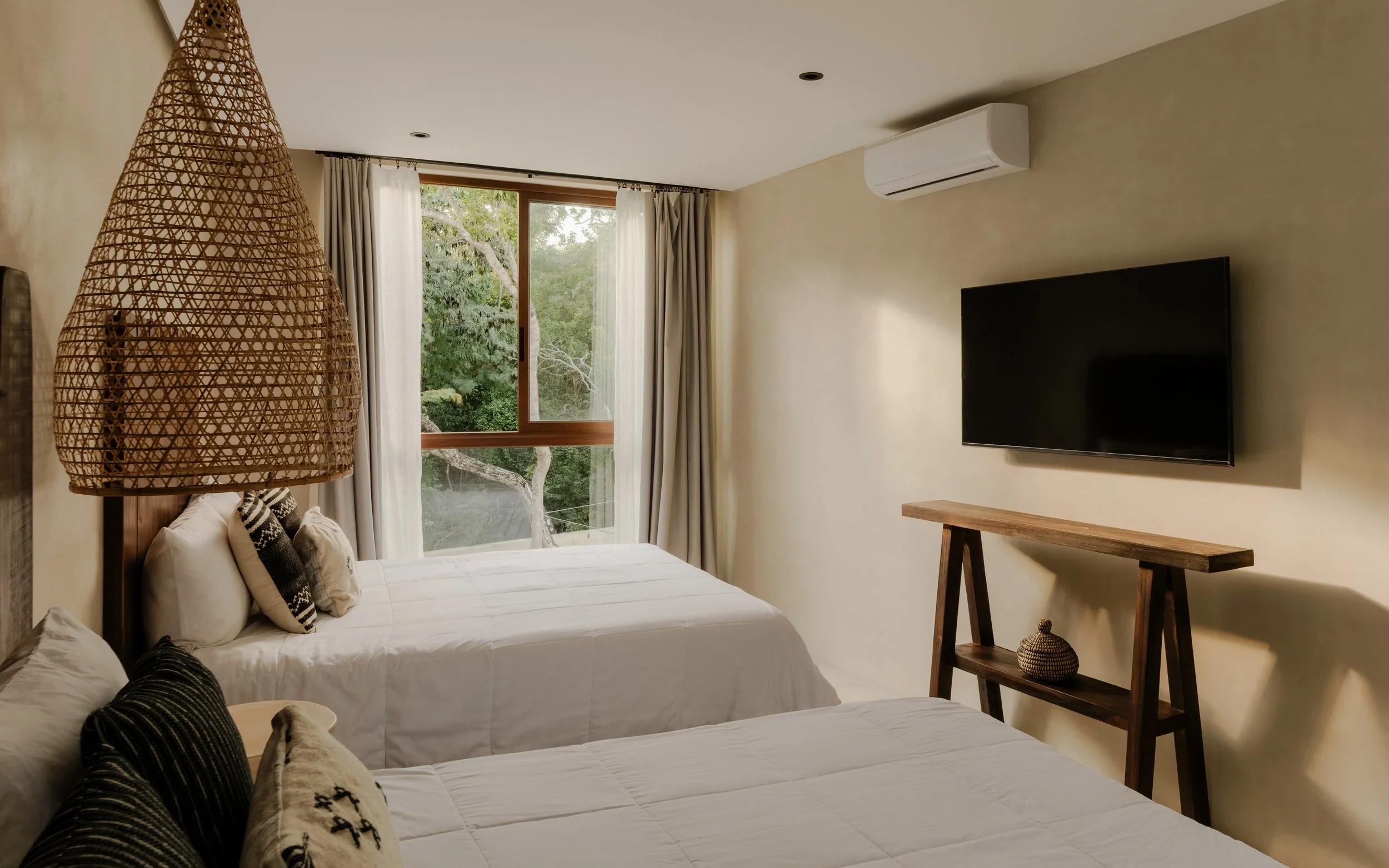 A bedroom with two beds, a large window with curtains showing trees outside, a wall-mounted TV, and a woven hanging light fixture.