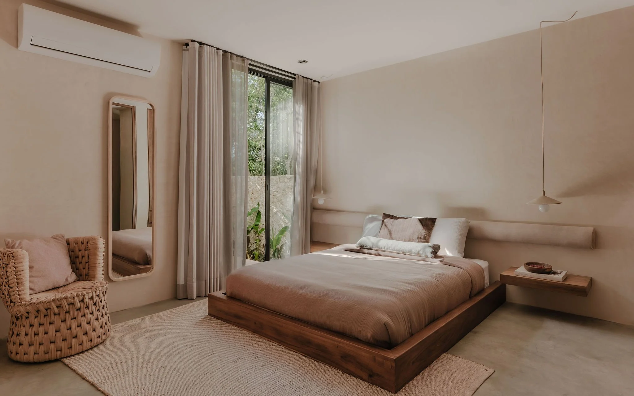 A bedroom with a bed, wicker chair, and large window with curtains, natural greenery outside, and minimal decor.