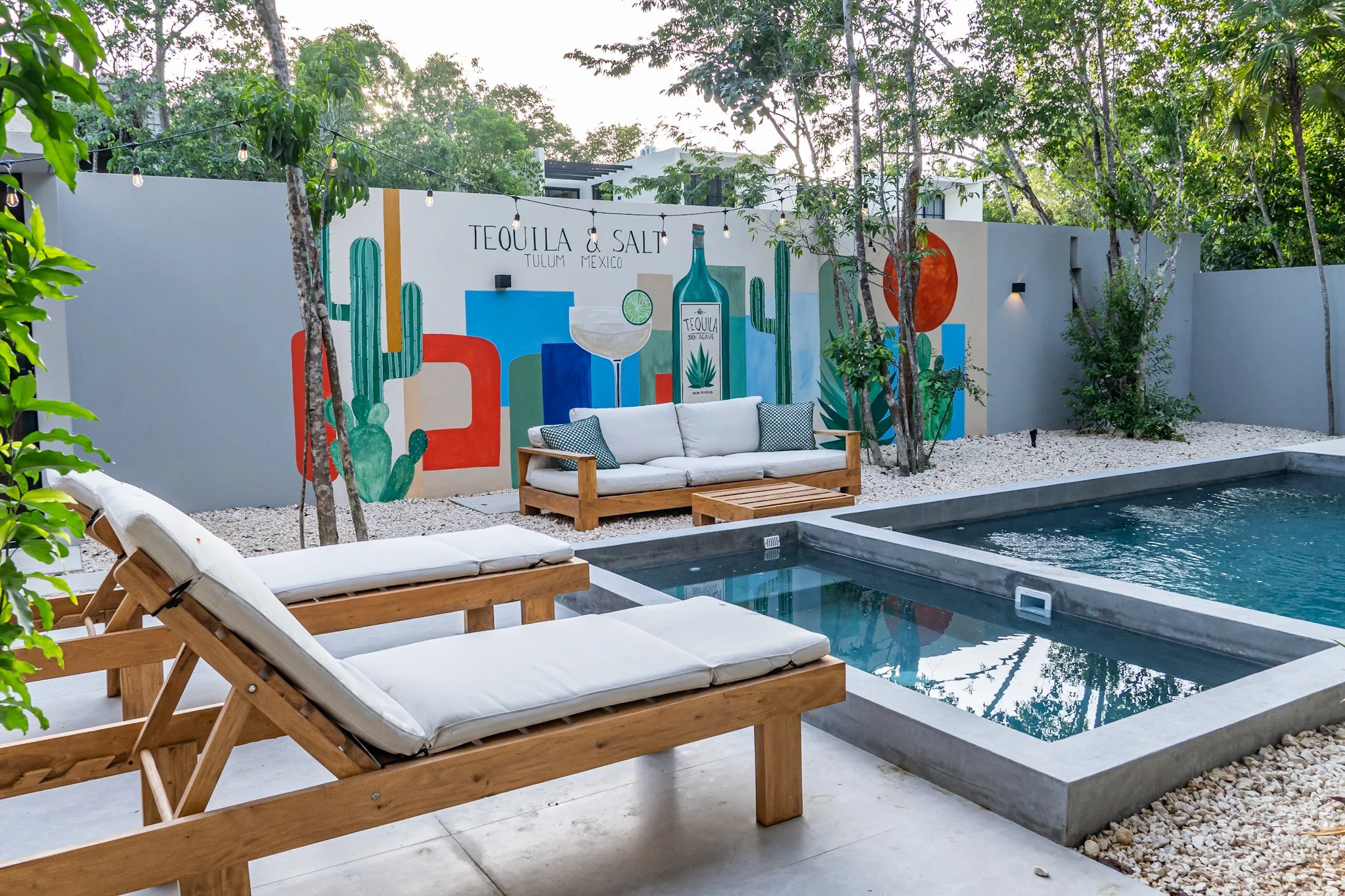 Outdoor lounge area with white cushioned chairs and wooden loungers beside a pool, a mural with desert cacti, and string lights hanging above, surrounded by trees.
