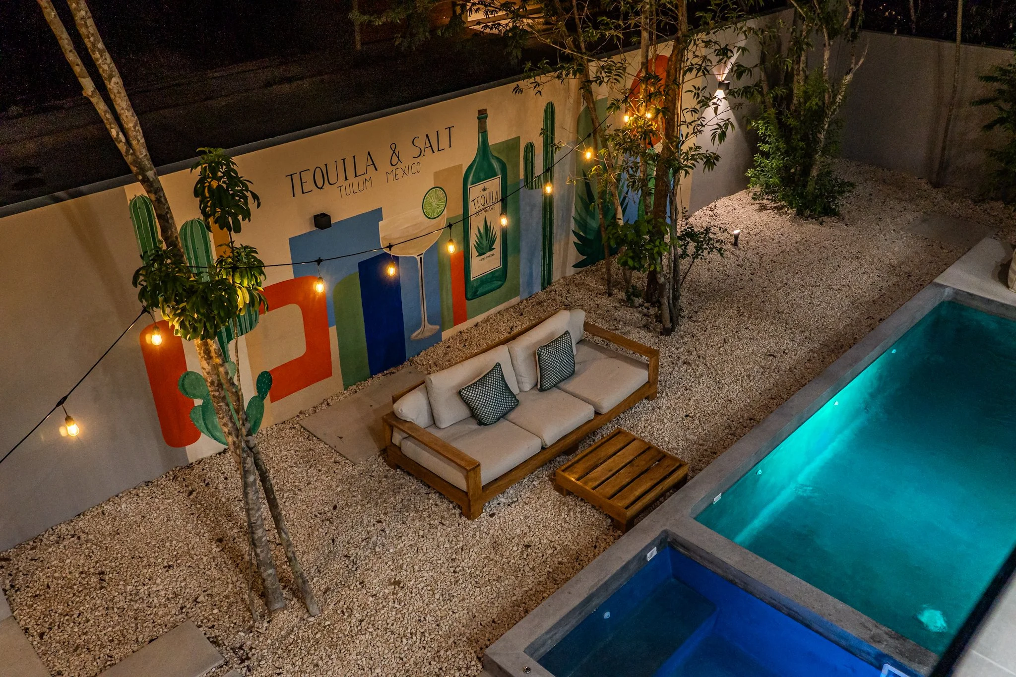 Nighttime view of an outdoor patio with a pool, seating area with a sofa and small table, decorative string lights, trees, a large mural with cactus and tequila bottles, and gravel ground.