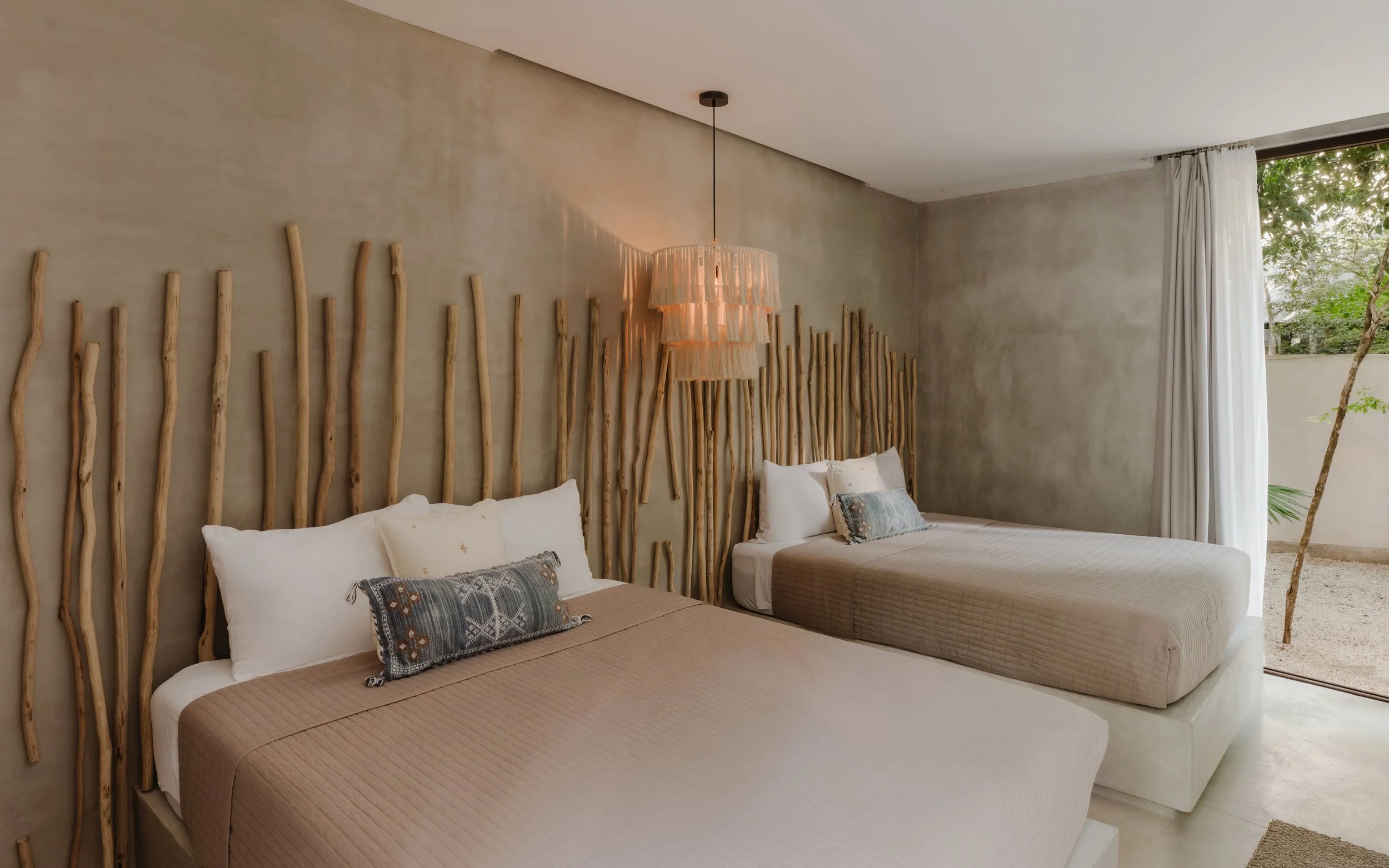 Two beds with beige quilts, decorated with pillows, in a bedroom with a rustic wood headboard, beige walls, a pendant lamp, and a large window with white curtains and a view of greenery outside.