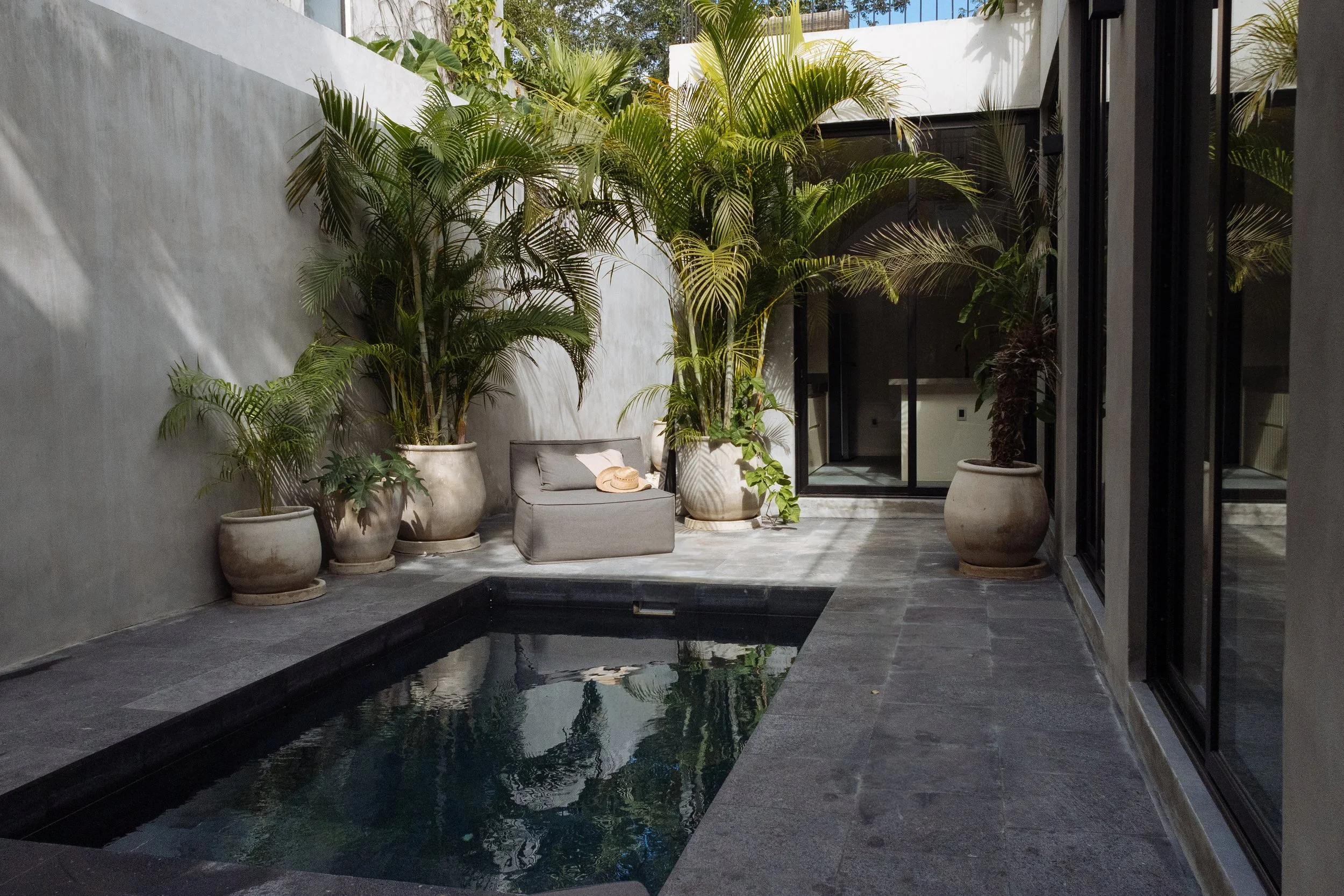 Indoor patio with a small rectangular pool, surrounded by potted palm trees and modern furniture, with large glass sliding doors and a white exterior wall.