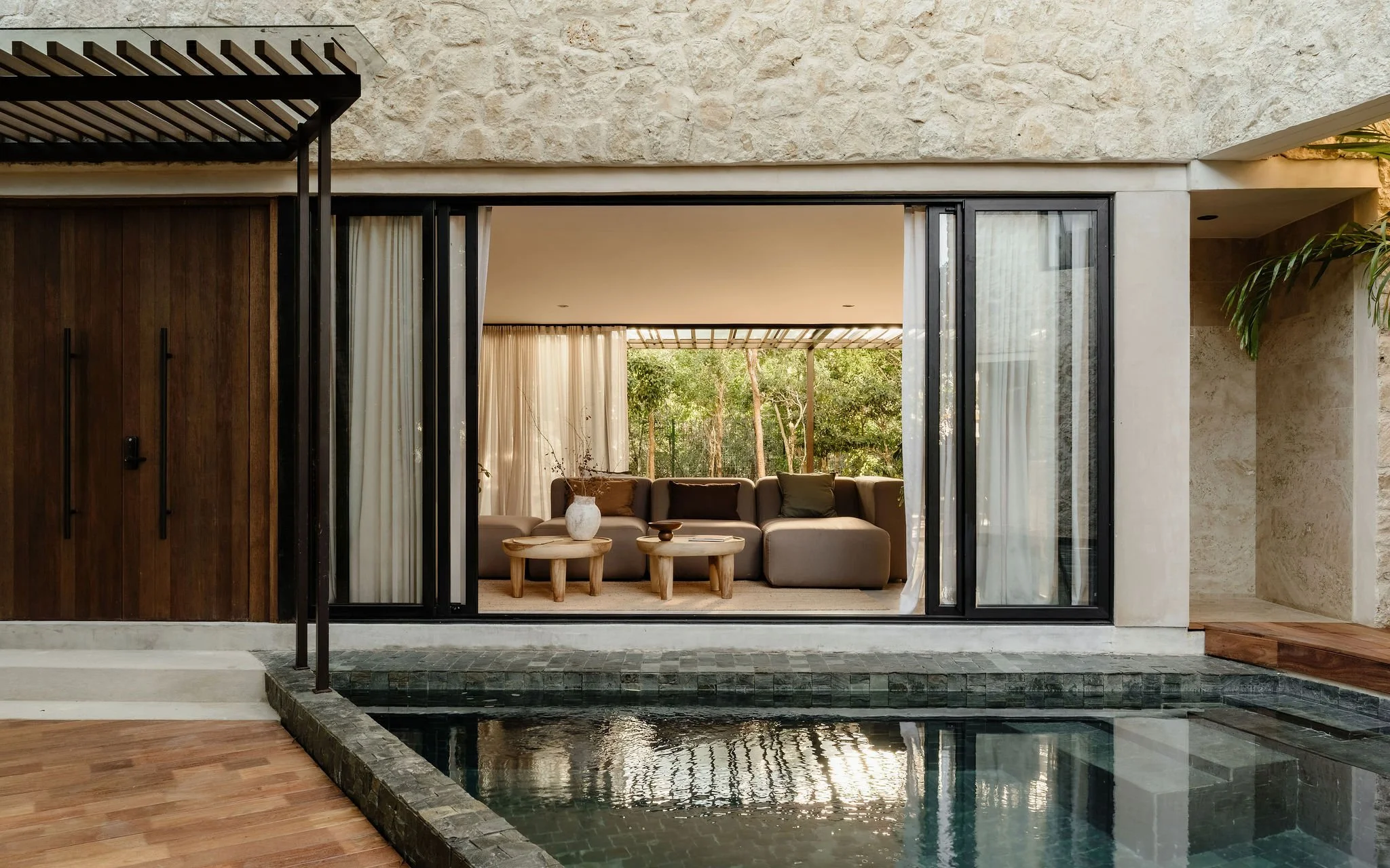 Living room with large glass sliding doors, beige sofa with cushions, potted plant, curtains, and outdoor greenery visible through the doors. Features a water feature or small pool in the foreground with stone border and wooden floor extending from the interior.