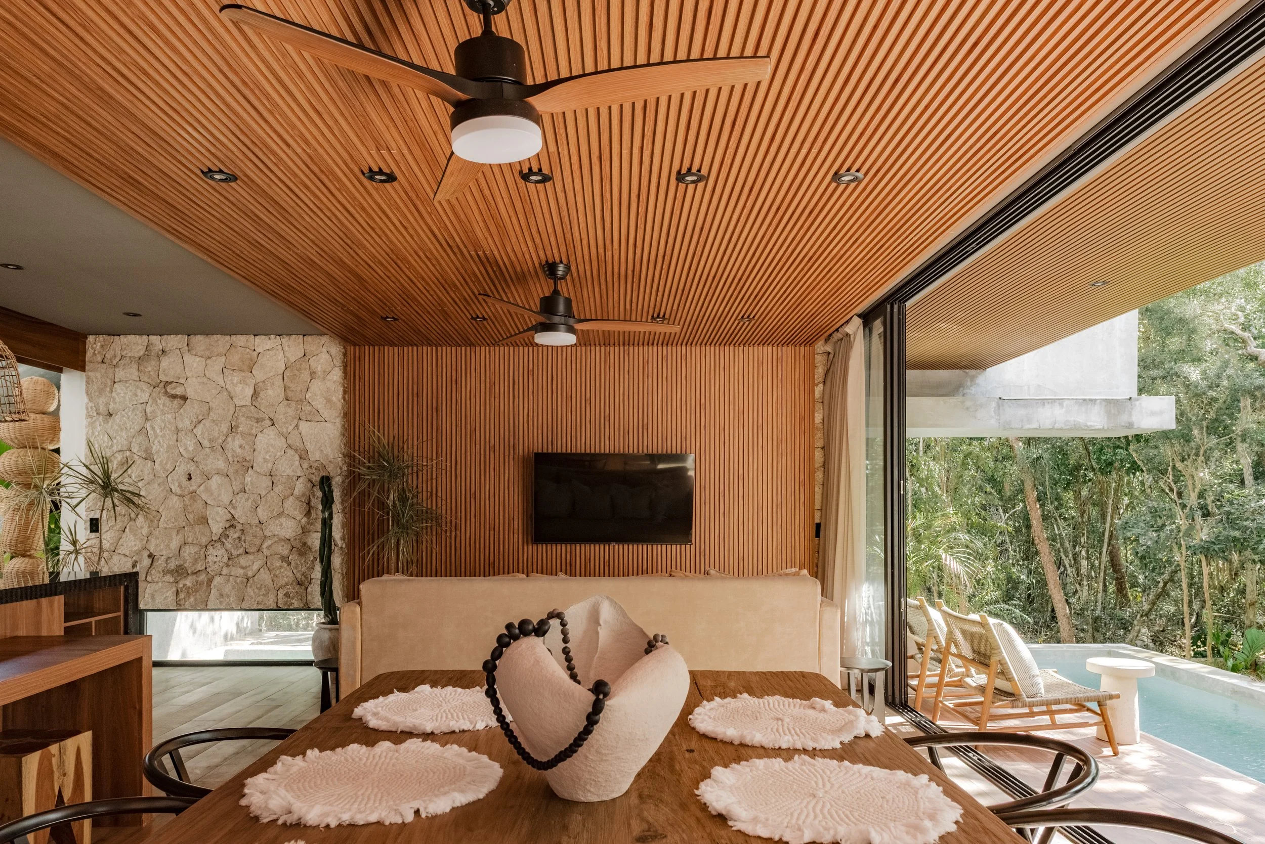 Living room with wooden ceiling fans, a stone wall, a large window with outdoor greenery, a beige sofa, and a wooden dining table with white cloth placemats and decorative sculpture.