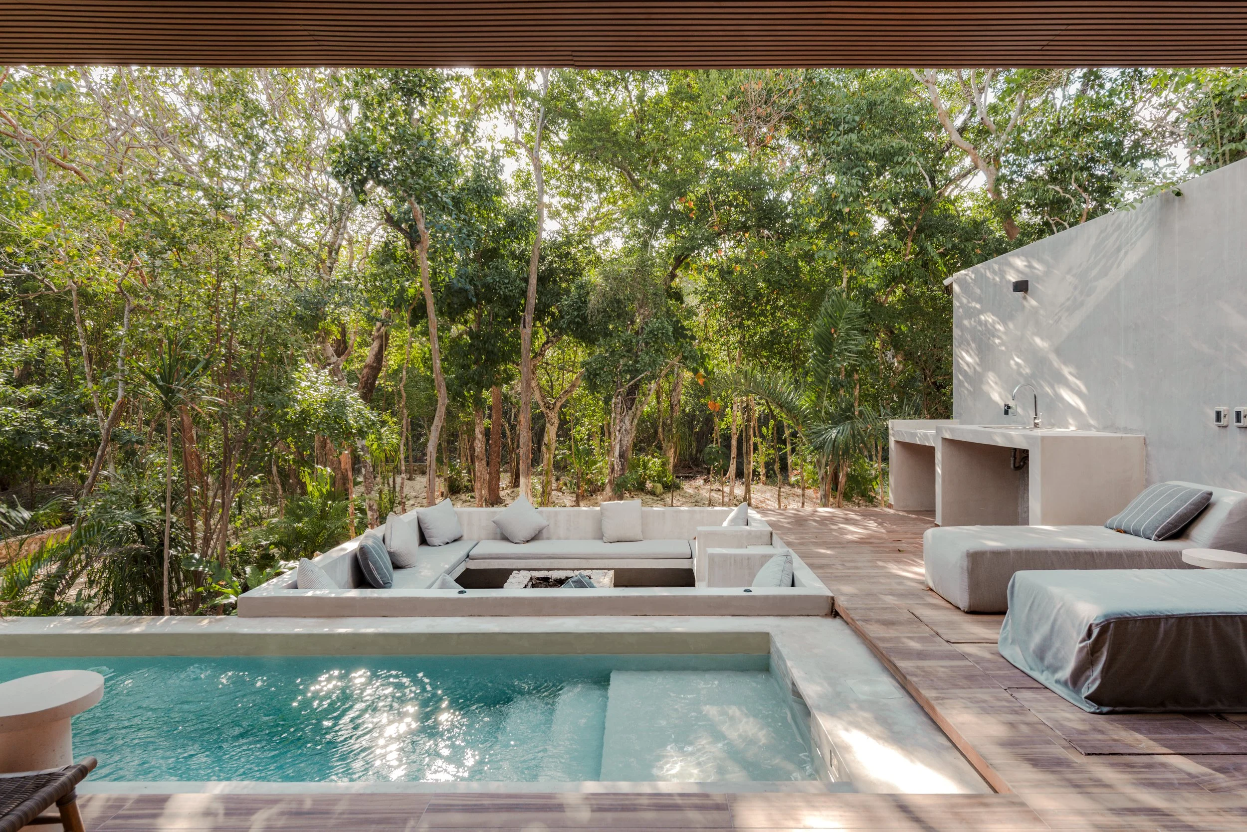 A modern outdoor patio with a swimming pool, surrounded by lush green trees and dense foliage, featuring white outdoor furniture and a fire pit.