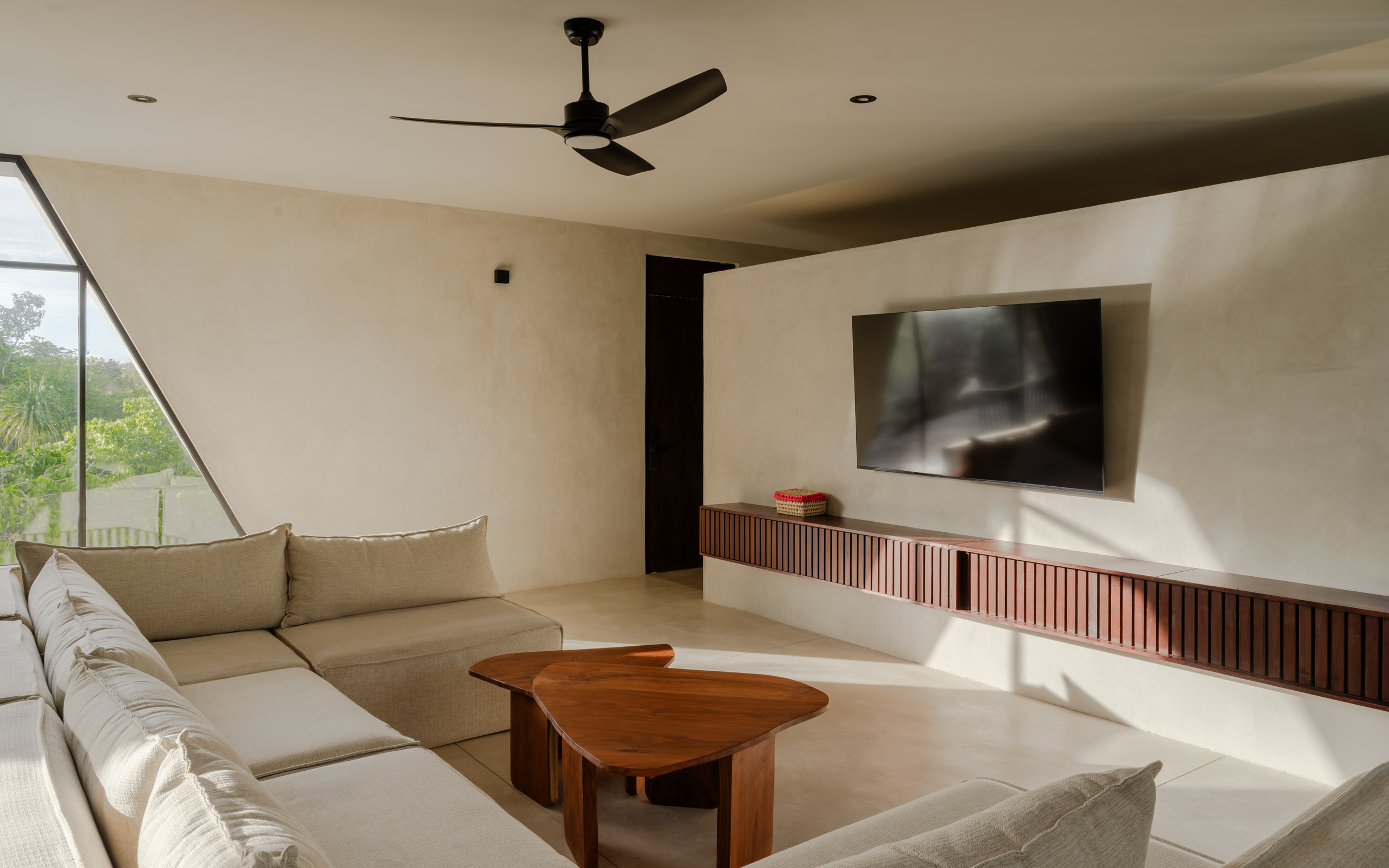 Living room with a beige sectional sofa, a wooden coffee table, a wall-mounted TV, and a large window with trees outside.