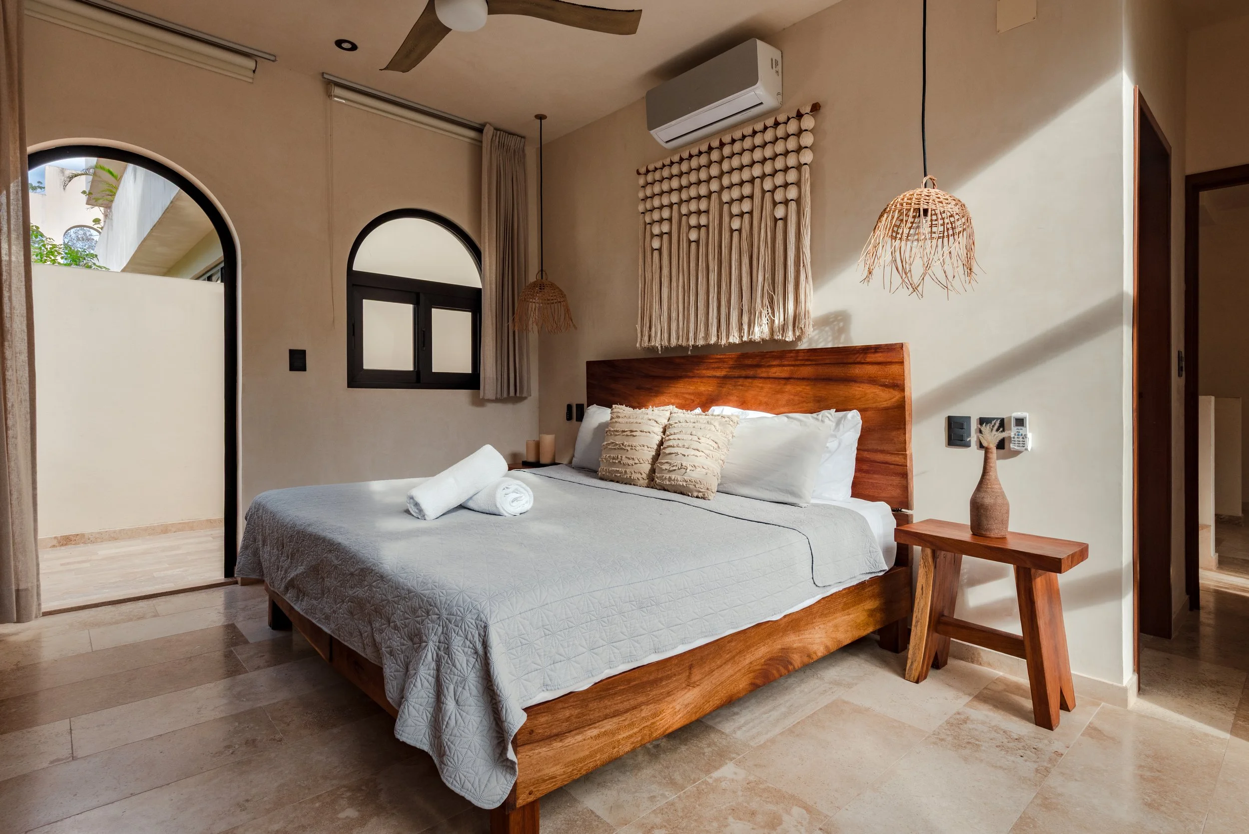 A bedroom with a wooden bed, two pillows, a white bedspread, and a rolled towel. There are two pendant lights, a wall decoration, an air conditioning unit, and a window with curtains. The room has beige walls and light-colored flooring.