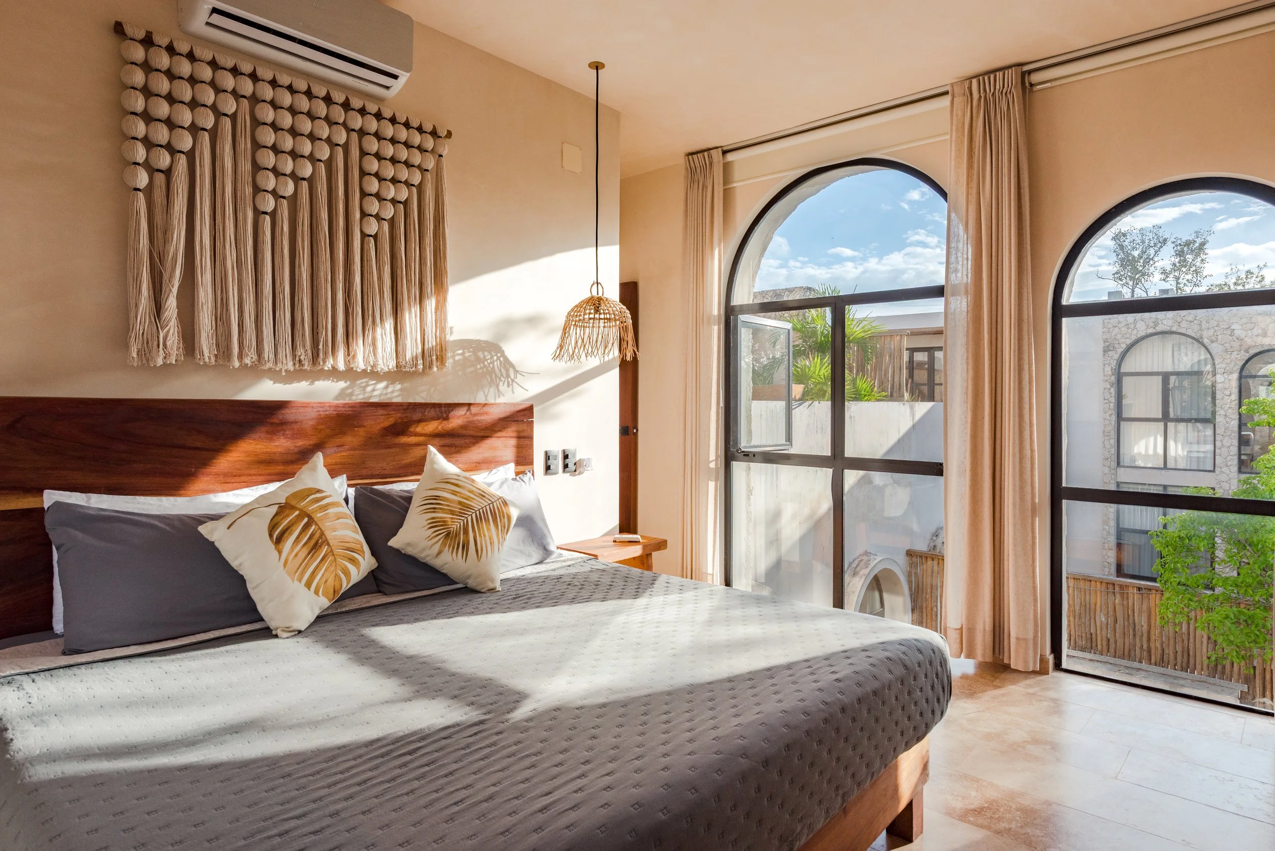 A bedroom with a bed, decorative pillows with leaf patterns, beige curtains, large arched windows showing outdoor buildings and greenery, and a wall hanging made of round wooden beads with tassels. Sunlight streams into the room.