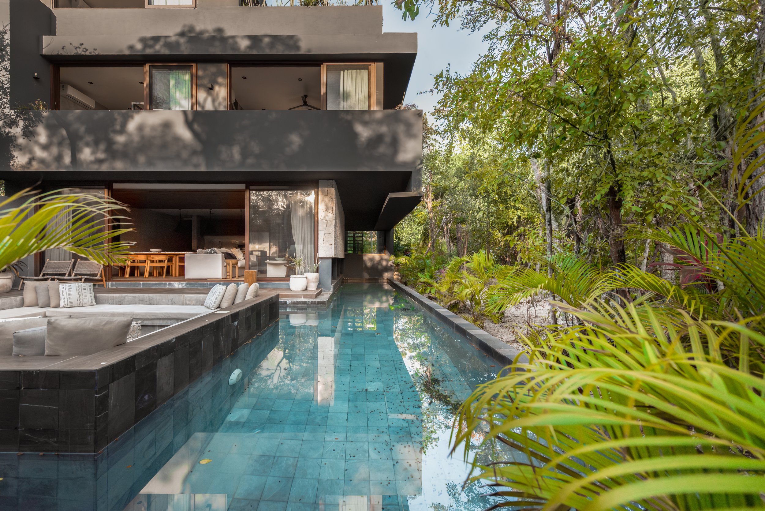 Modern multi-story house with outdoor pool, surrounded by lush green trees and plants.