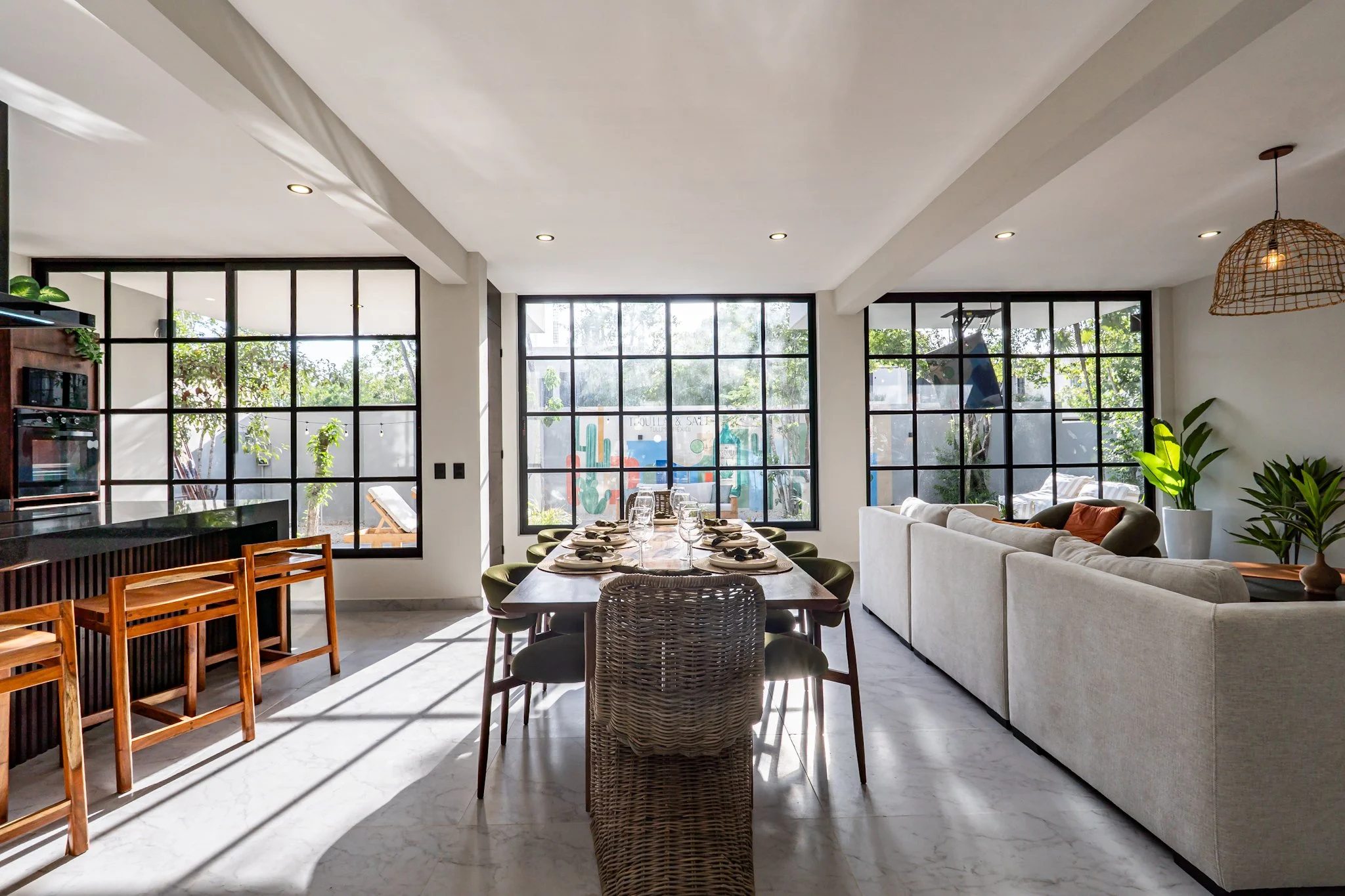Modern dining and living area with large windows allowing natural light, a wooden dining table set with glassware, and a white sofa with cushions, surrounded by indoor plants and warm lighting.