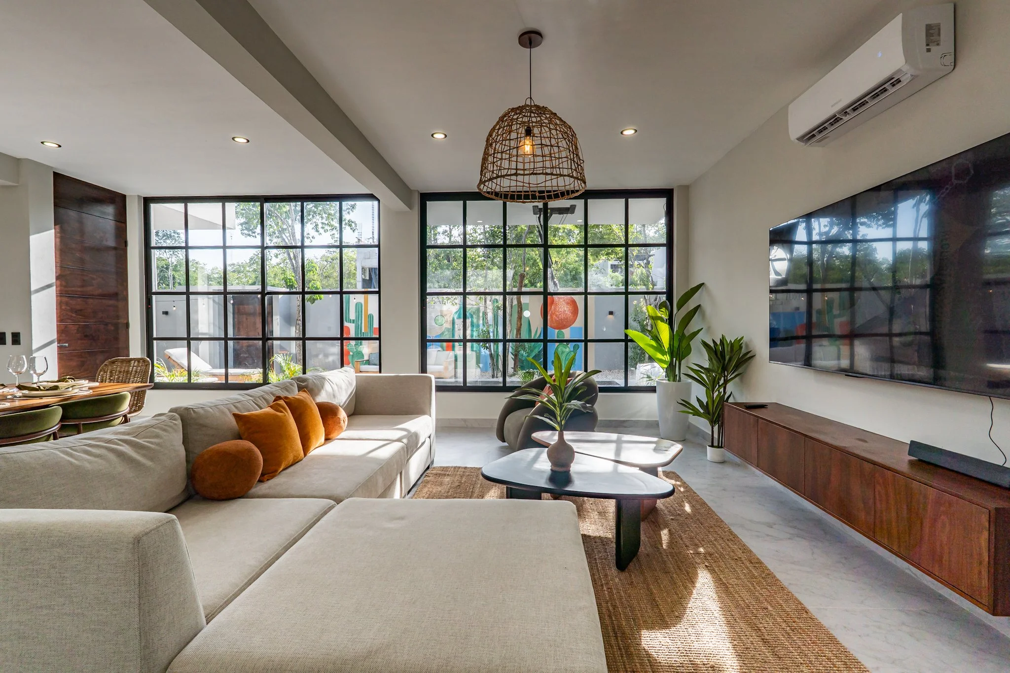 Bright living room with large black-framed windows, a white sectional sofa with orange and beige cushions, a wooden coffee table with a white vase, indoor plants, a wall-mounted TV, a woven pendant light, and an air conditioning unit on the ceiling.