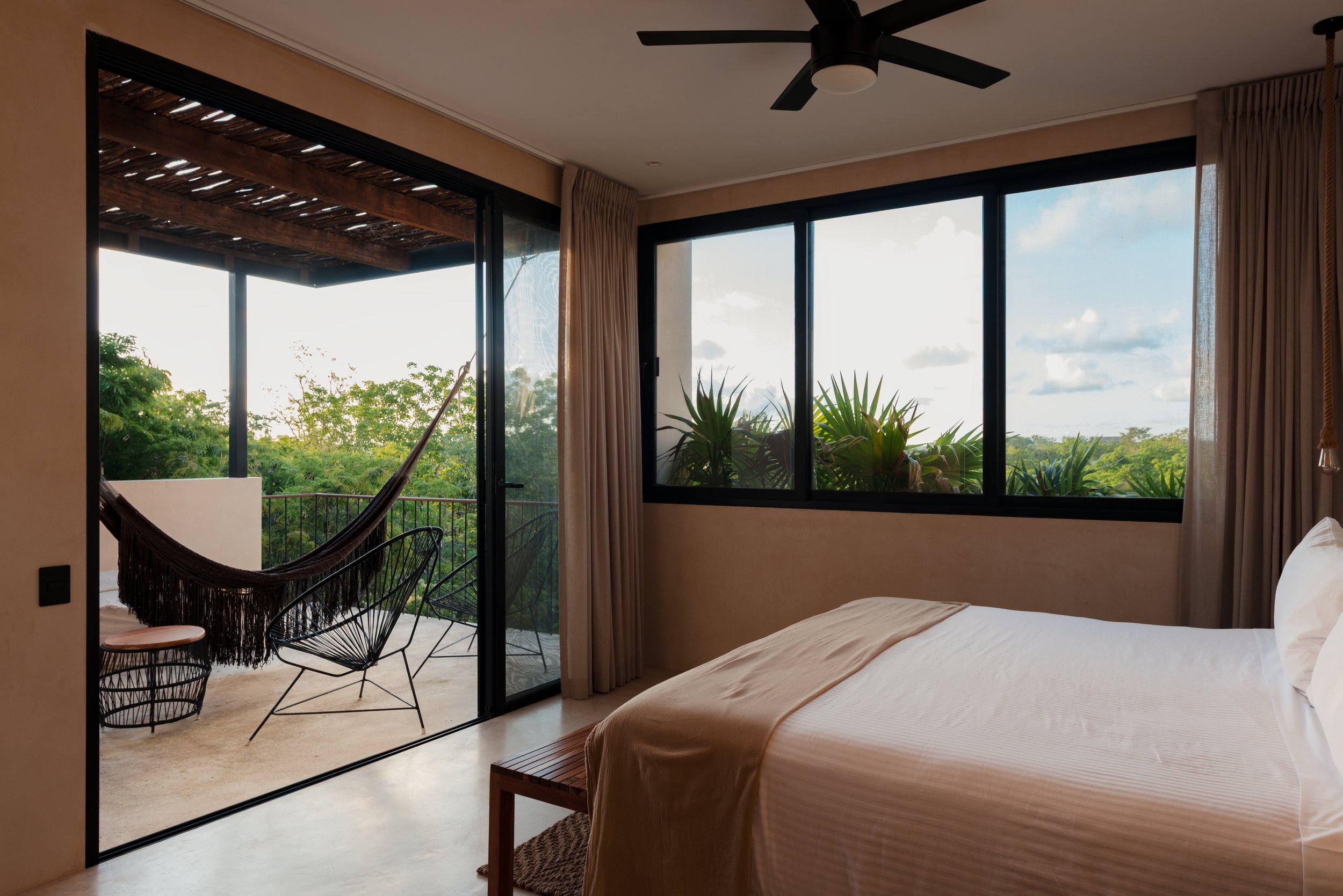 A bedroom with a large window showing lush green trees outside, a bed with a beige bedspread, a wooden bench at the foot of the bed, and a balcony with black chairs and a hammock. Curtains frame the window and a ceiling fan is on the ceiling.