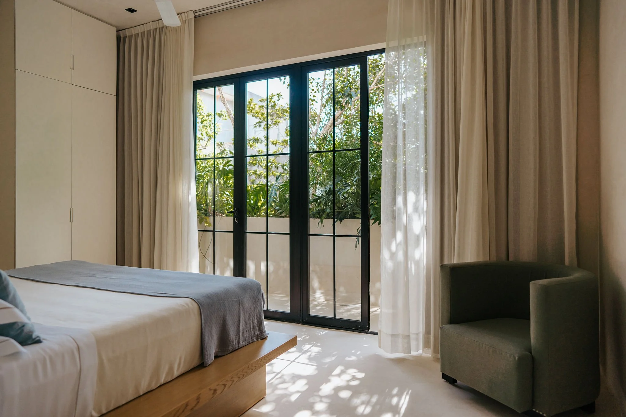 A bedroom with a large window and glass door overlooking greenery, a bed with white bedding, beige curtains, and a gray armchair.