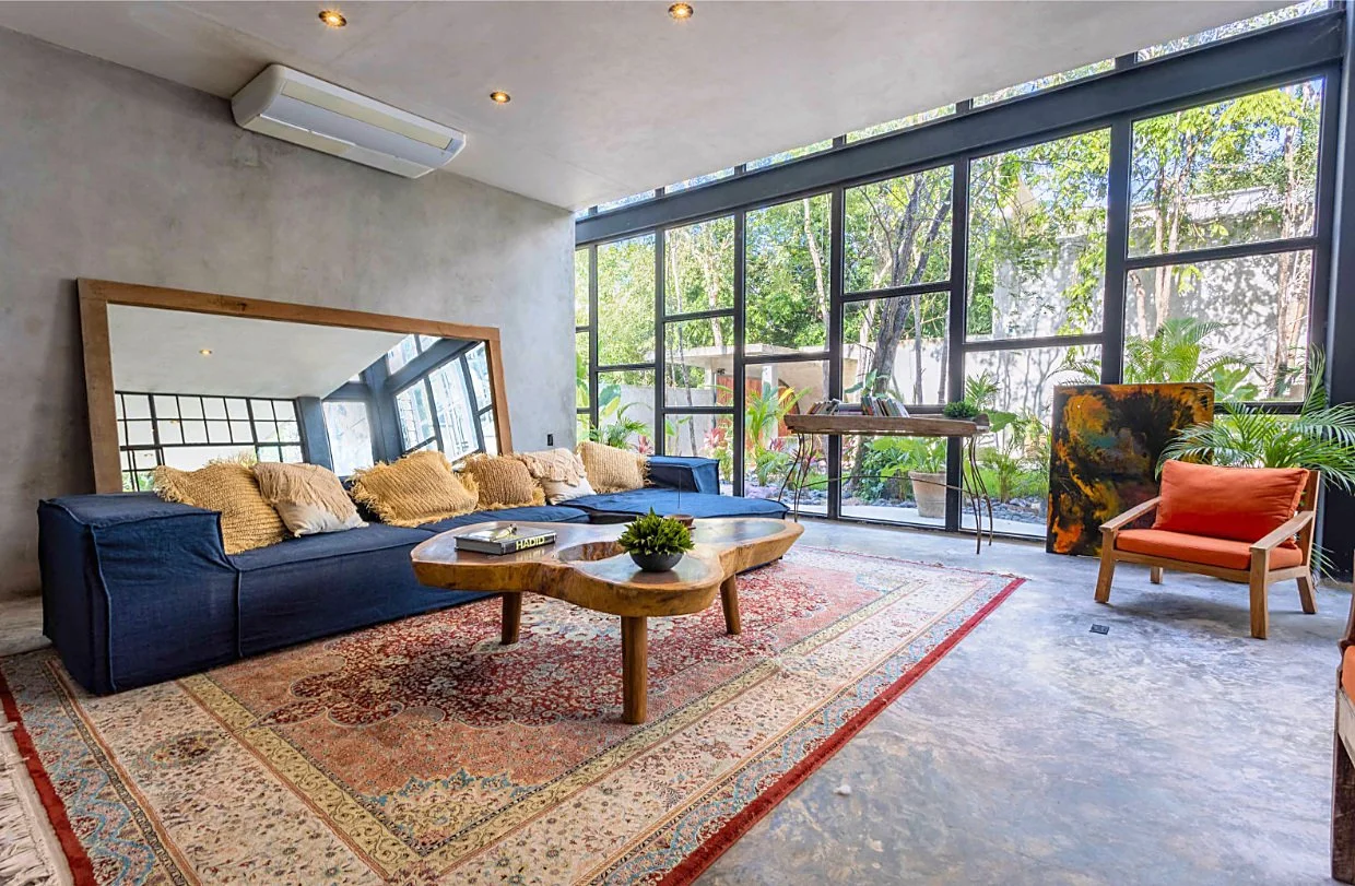 A modern living room with large window walls, a blue sofa with beige pillows, a wooden coffee table on an ornate rug, a large mirror leaning against the wall, and various plants and artwork near the windows.
