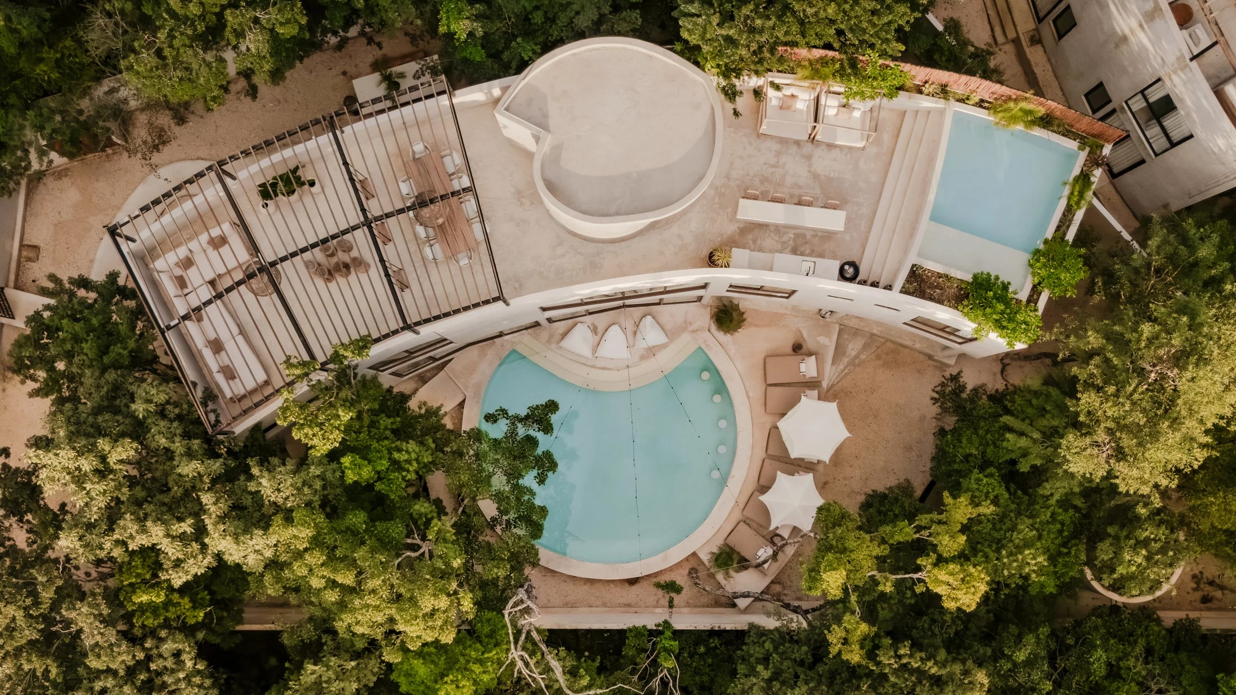 An aerial view of a modern, multi-story hotel with a rooftop pool, surrounded by lush green trees and landscaped outdoor areas, including lounge chairs and umbrellas.